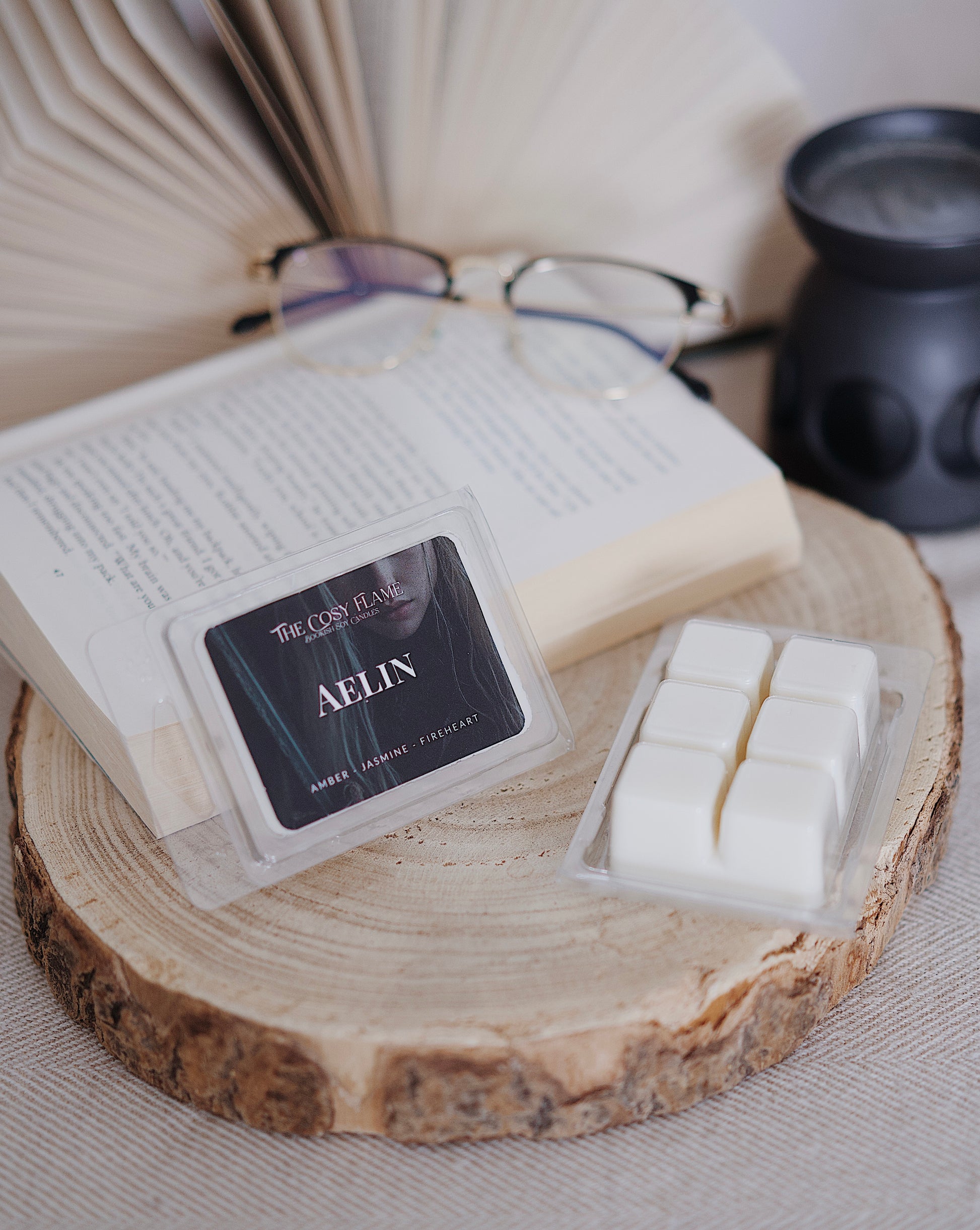 Aelin soy wax melts by The Cosy Flame, featuring amber and jasmine scent notes, displayed on a wooden slice beside an open book and glasses.