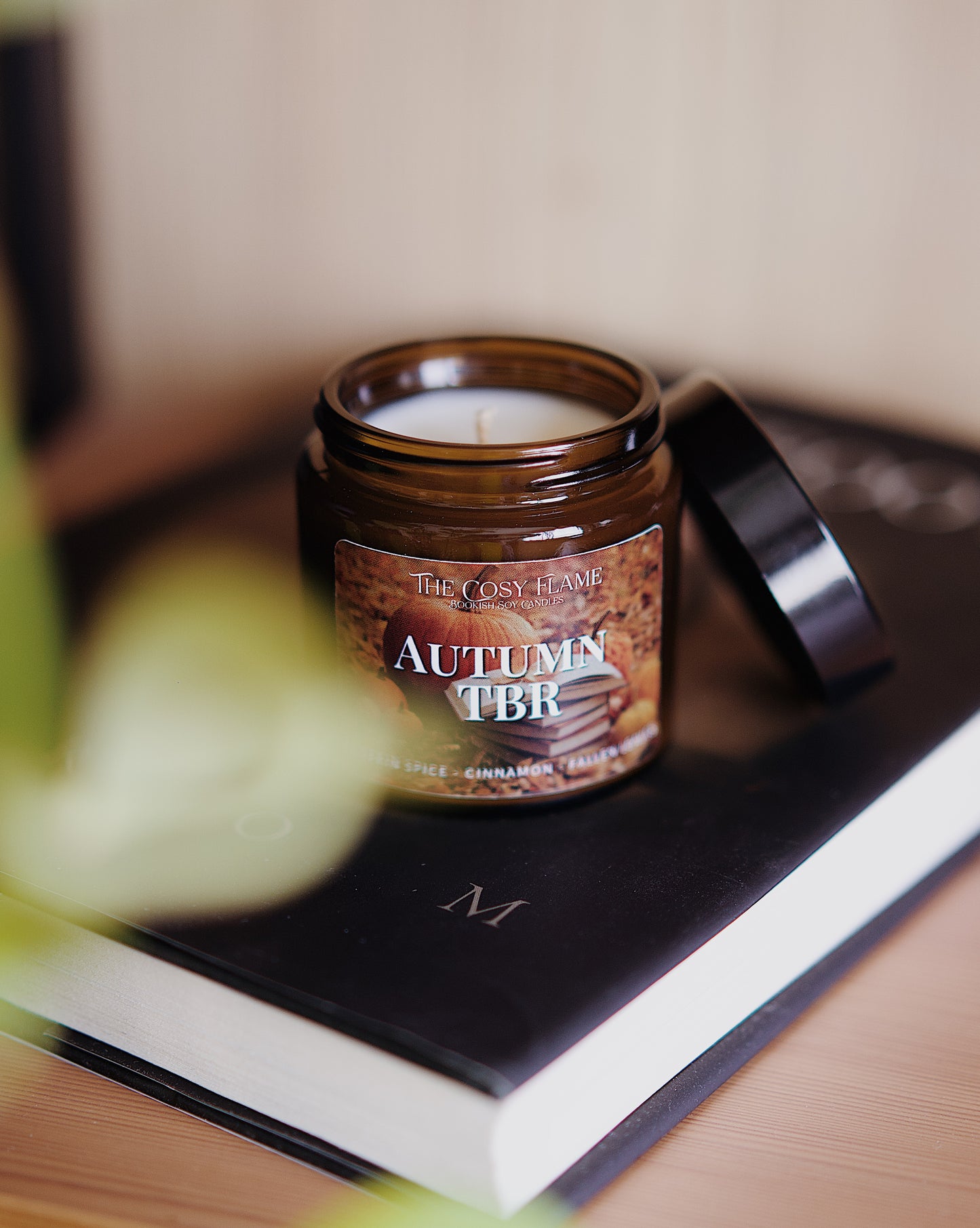 Amber glass candle jar labelled ‘Autumn TBR’ from The Cosy Flame, scented with pumpkin spice and cinnamon, and a book in the background.