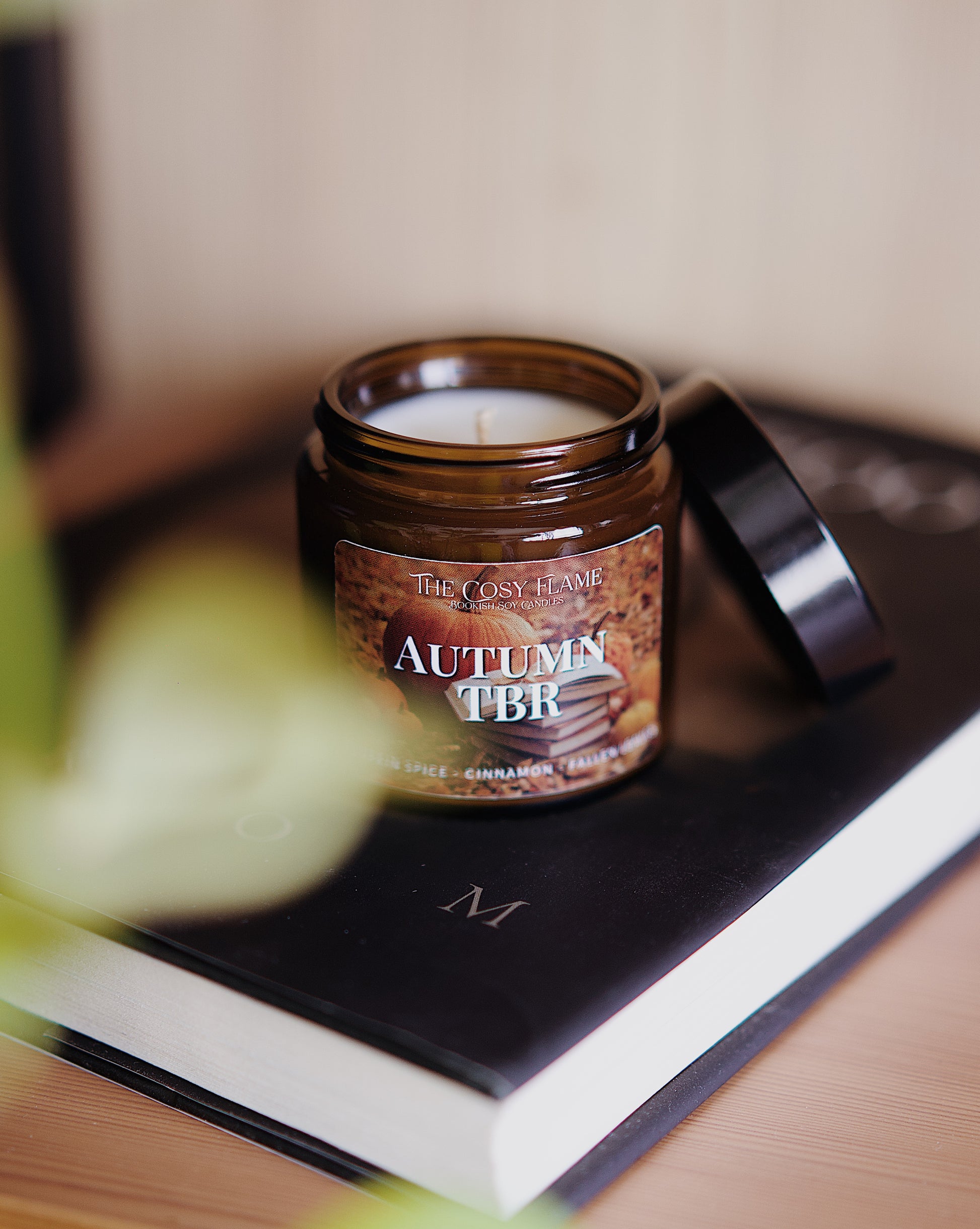 Amber glass candle jar labelled ‘Autumn TBR’ from The Cosy Flame, scented with pumpkin spice and cinnamon, and a book in the background.