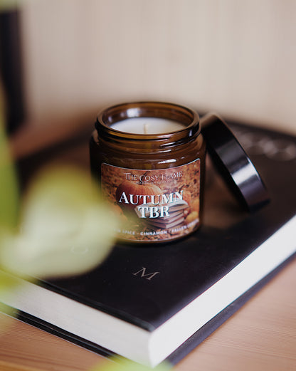 Amber glass candle jar labelled ‘Autumn TBR’ from The Cosy Flame, scented with pumpkin spice and cinnamon, and a book in the background.