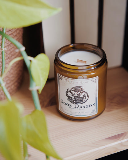 Book Dragon soy candle by The Cosy Flame in an amber glass jar with a wooden wick, placed on a wooden shelf. Label features a sleeping dragon illustration and featuring the scents orange and amber.