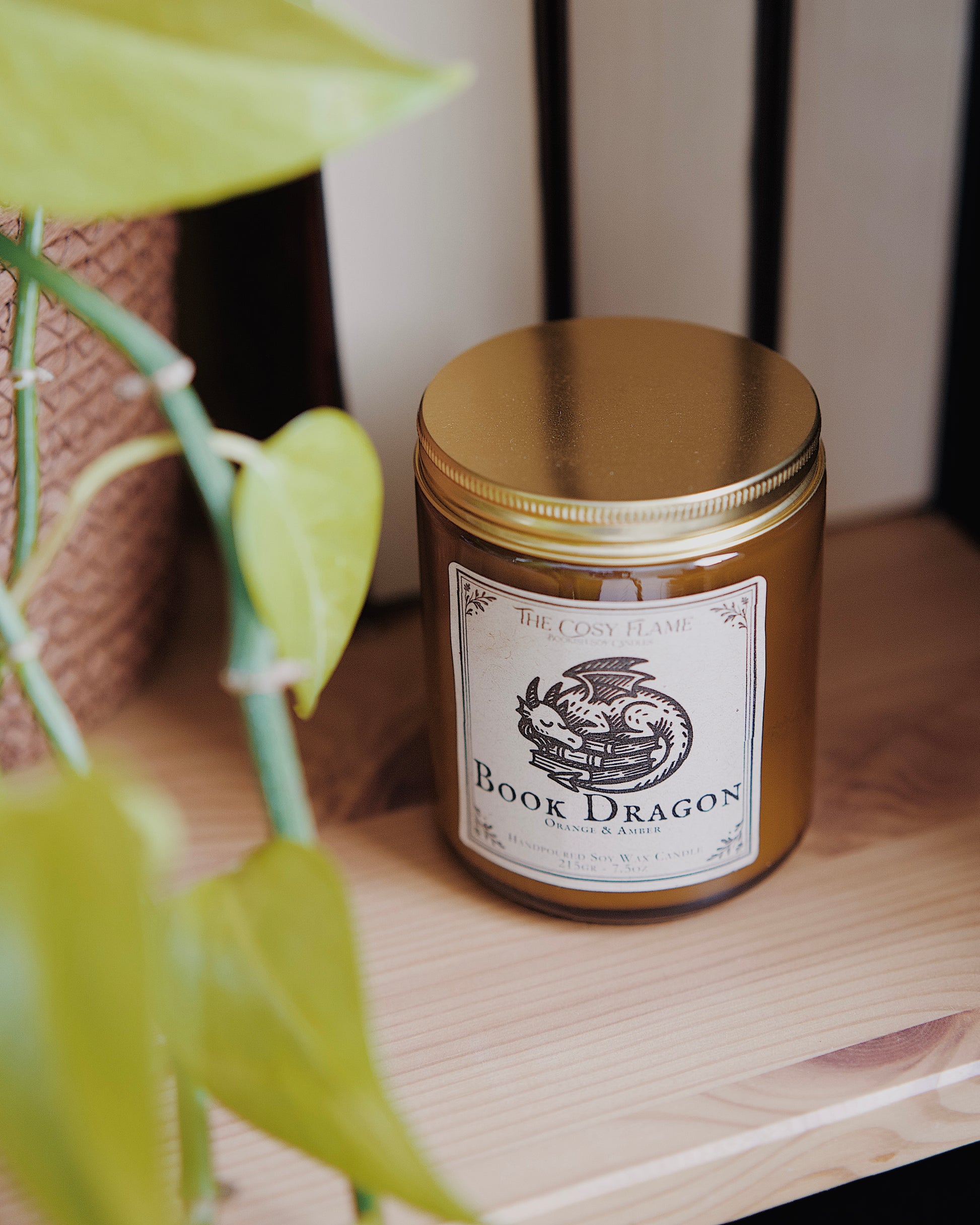 Book Dragon soy candle by The Cosy Flame in an amber glass jar with a golden lid, placed on a wooden shelf. Label features a sleeping dragon illustration and featuring the scents orange and amber.
