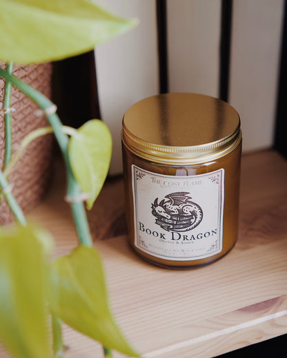 Book Dragon soy candle by The Cosy Flame in an amber glass jar with a golden lid, placed on a wooden shelf. Label features a sleeping dragon illustration and featuring the scents orange and amber.