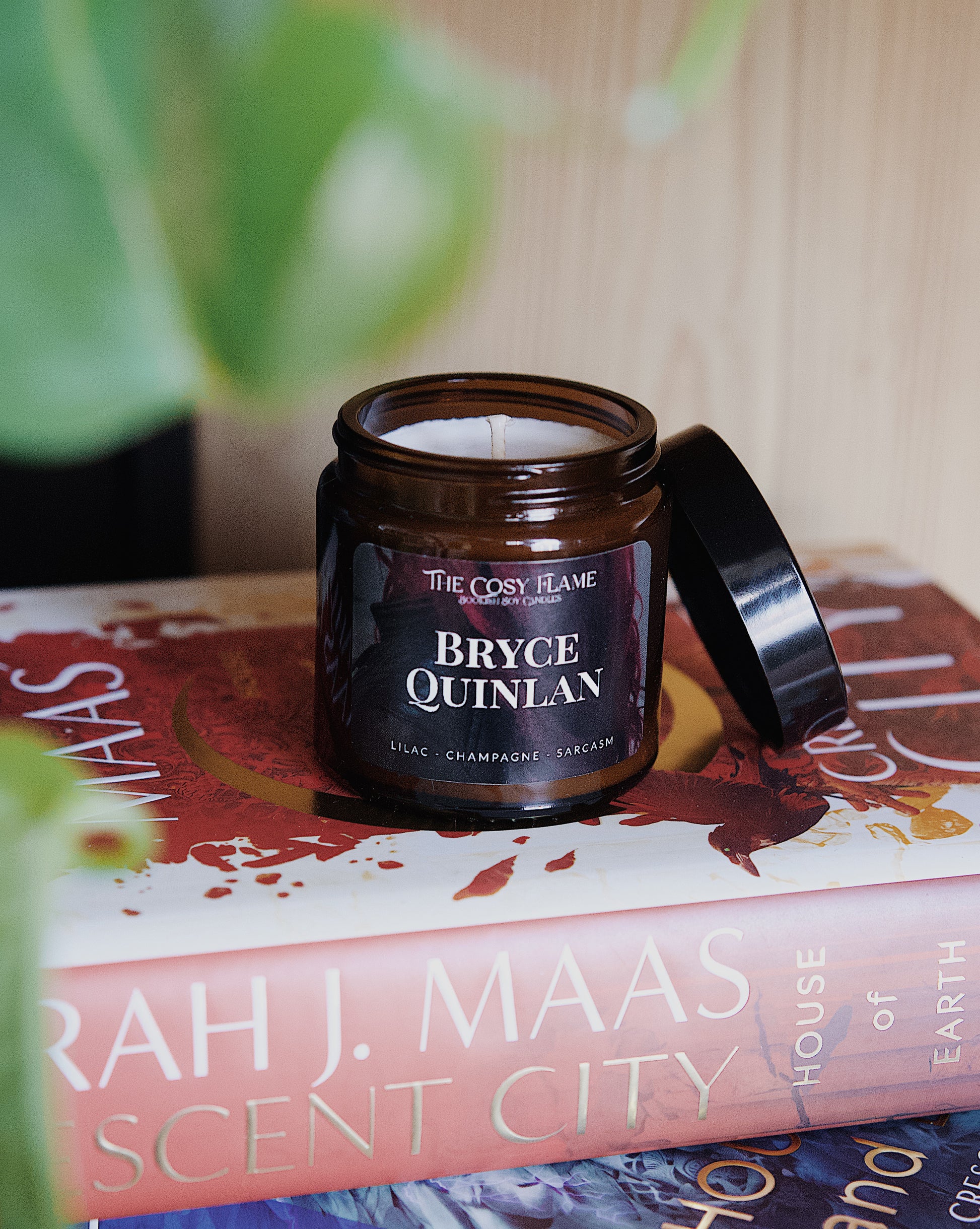 Amber glass candle jar labelled ‘Bryce Quinlan’ from The Cosy Flame, scented with lilac and champagne, and Crescent City books in the background.
