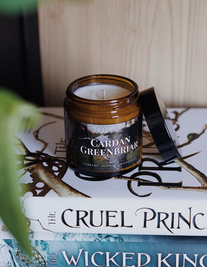 Amber glass candle jar labelled ‘Cardan Greenbriar’ from The Cosy Flame, scented with currant and almond, and The Cruel Prince books in the background.