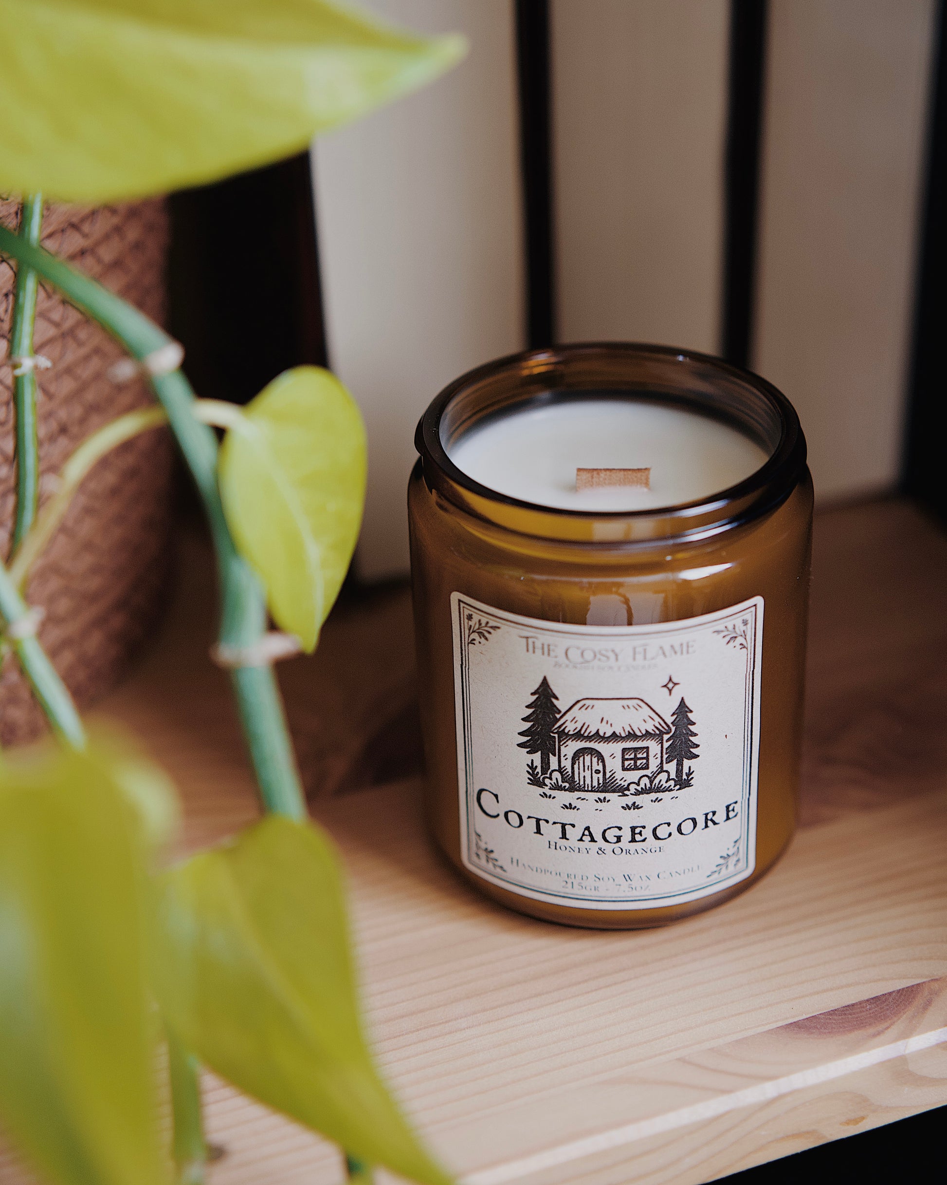 Cottagecore soy candle by The Cosy Flame in an amber glass jar with a wooden wick, placed on a wooden shelf. Label features a cosy cottage illustration and featuring the scents honey and orange.