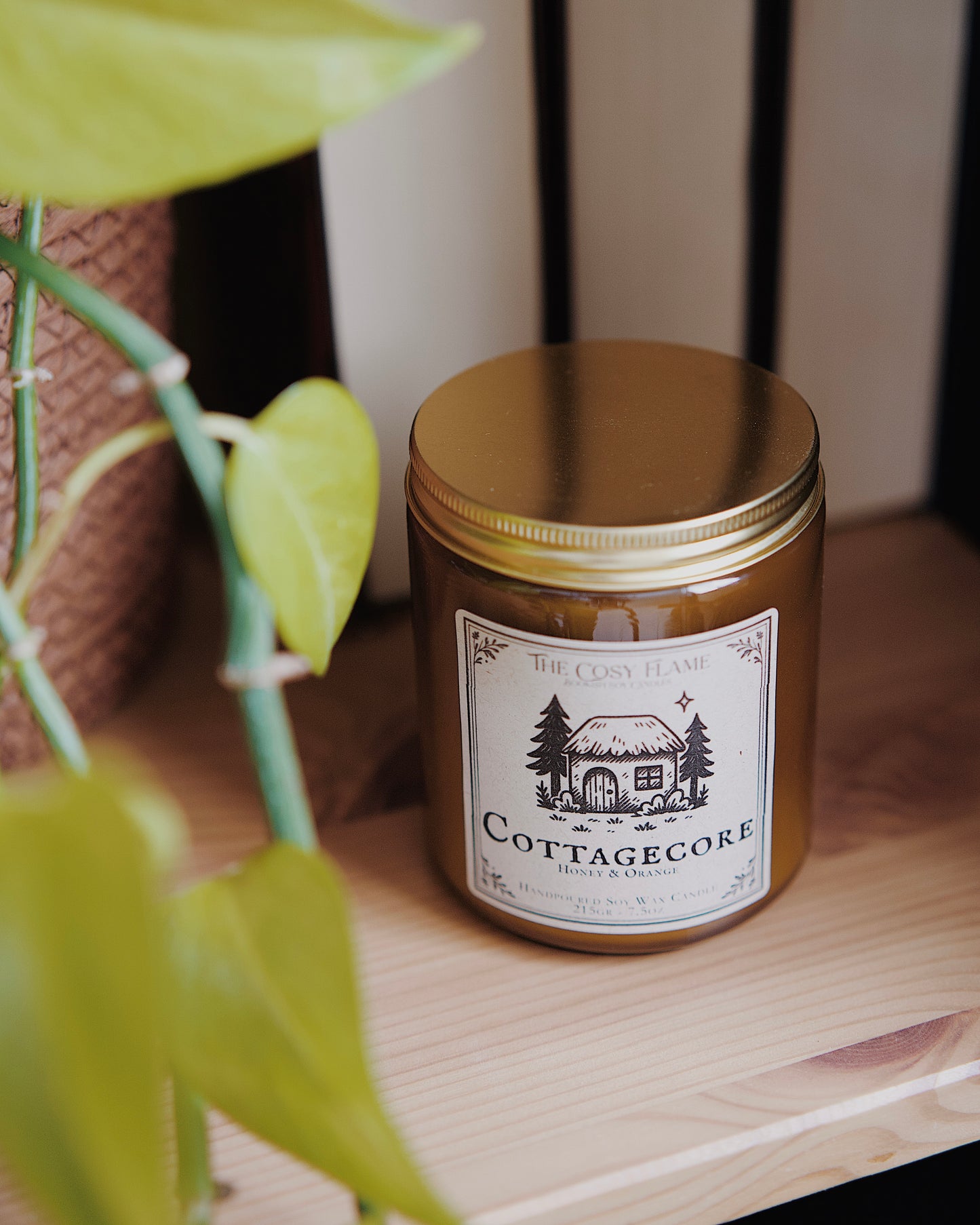 Cottagecore soy candle by The Cosy Flame in an amber glass jar with a golden lid, placed on a wooden shelf. Label features a cosy cottage illustration and featuring the scents honey and orange.