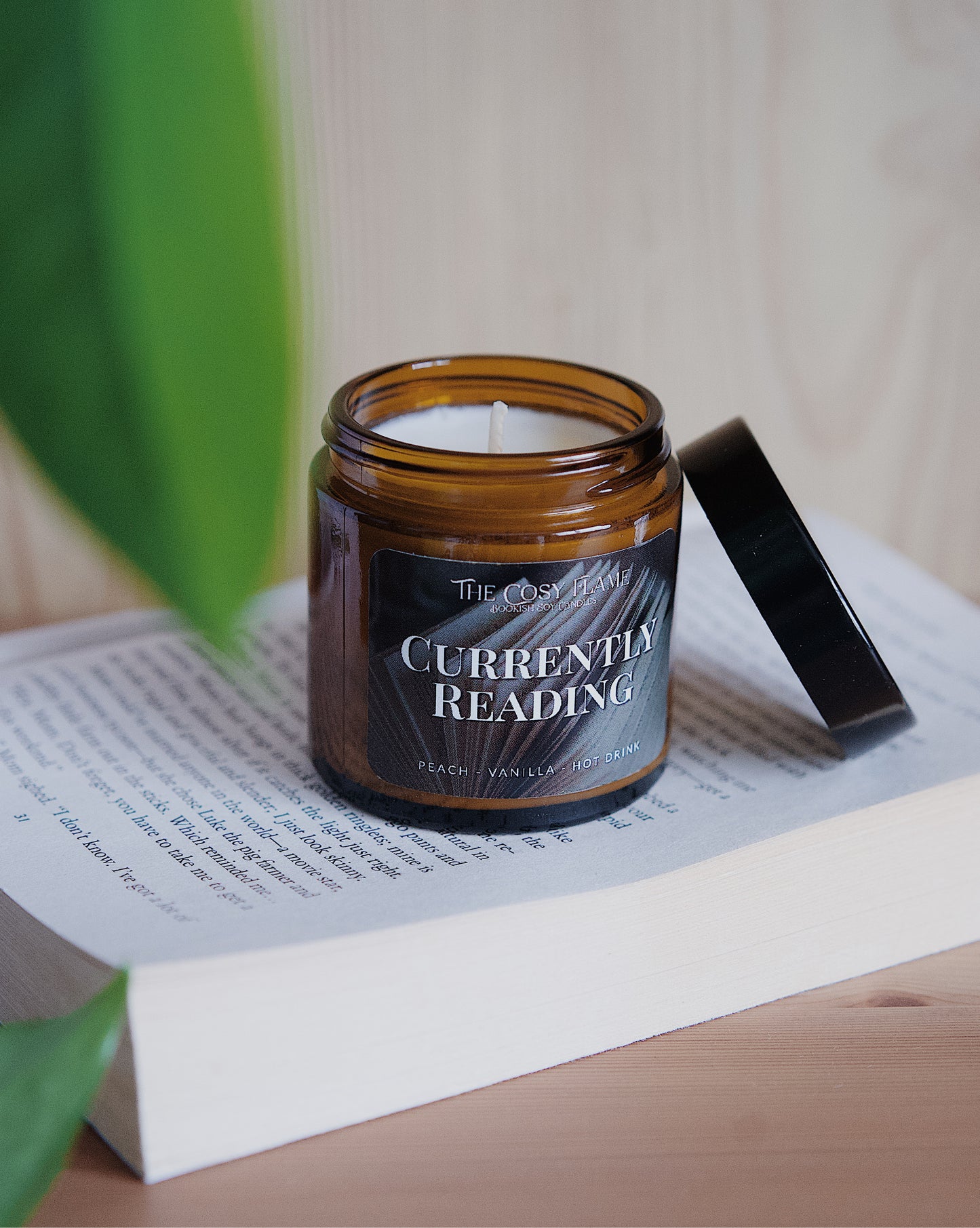 Amber glass candle jar labelled ‘Currently Reading’ from The Cosy Flame, scented with peach and vanilla, and a book in the background.