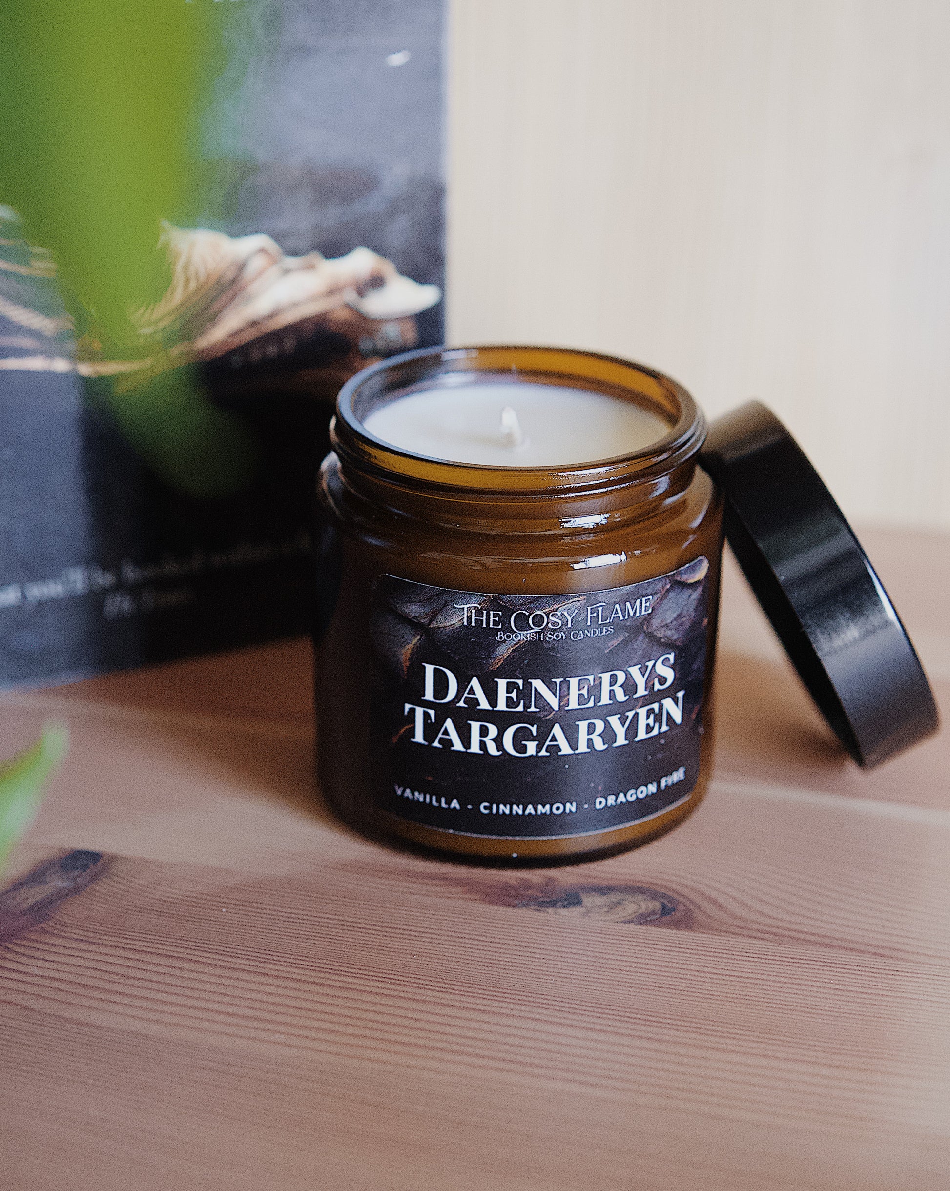 Amber glass candle jar labelled ‘Daenerys Targaryen’ from The Cosy Flame, scented with vanilla and cinnamon, and Game of Thrones book in the background.