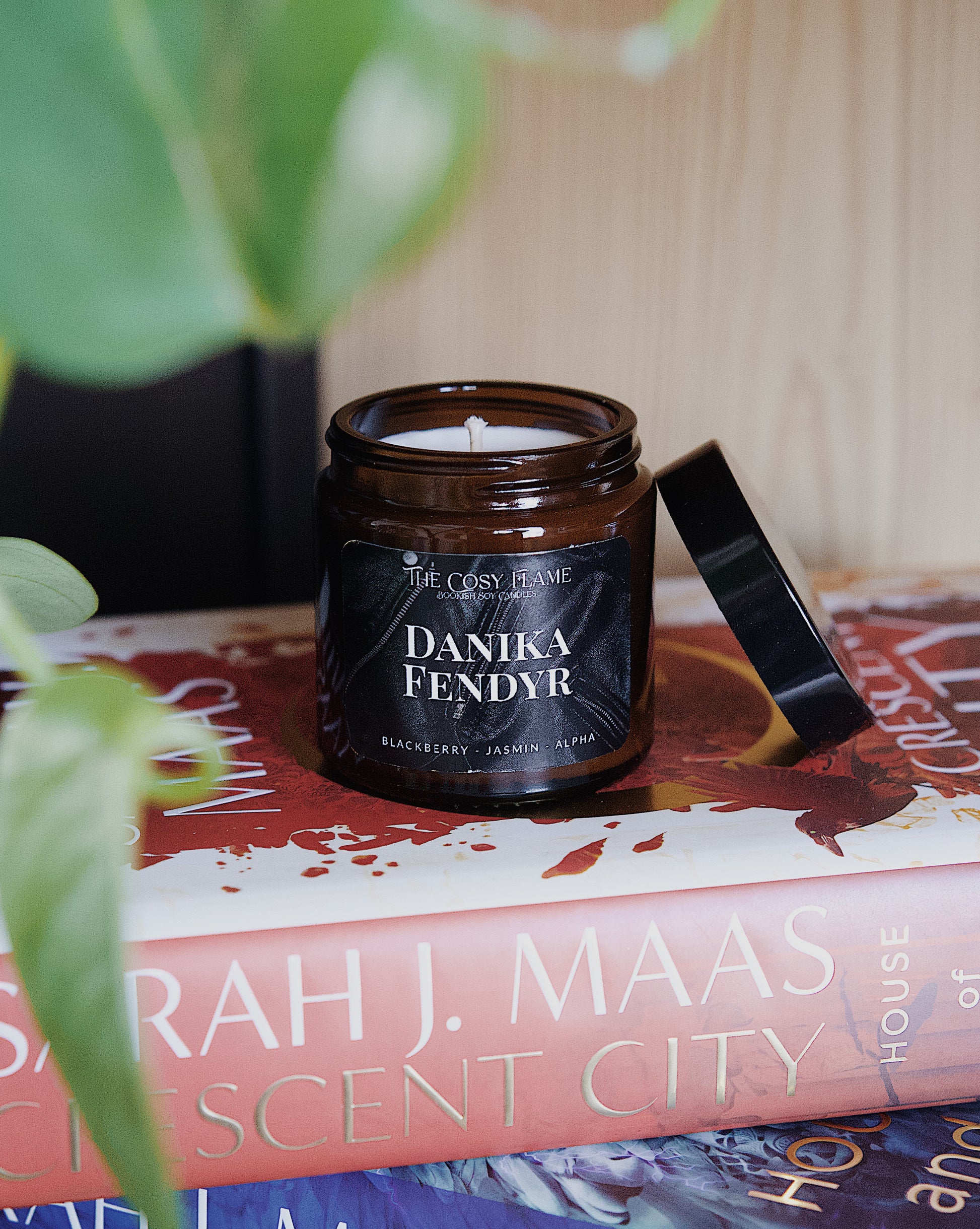 Amber glass candle jar labelled ‘Danika Fendyr’ from The Cosy Flame, scented with blackberry and jasmin, and Crescent City books in the background.