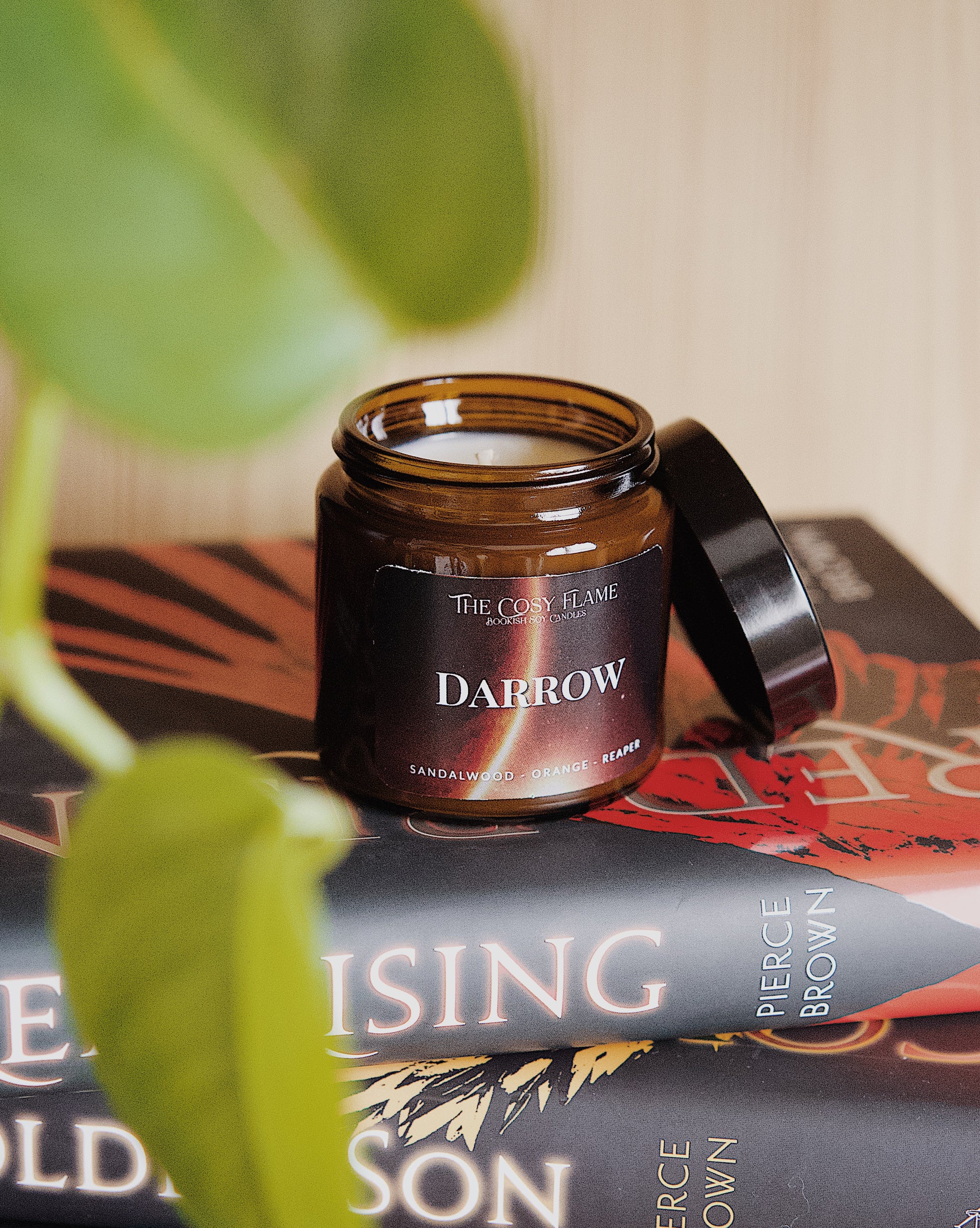 Amber glass candle jar labelled ‘Darrow’ from The Cosy Flame, scented with sandalwood and orange, and Red Rising books in the background.