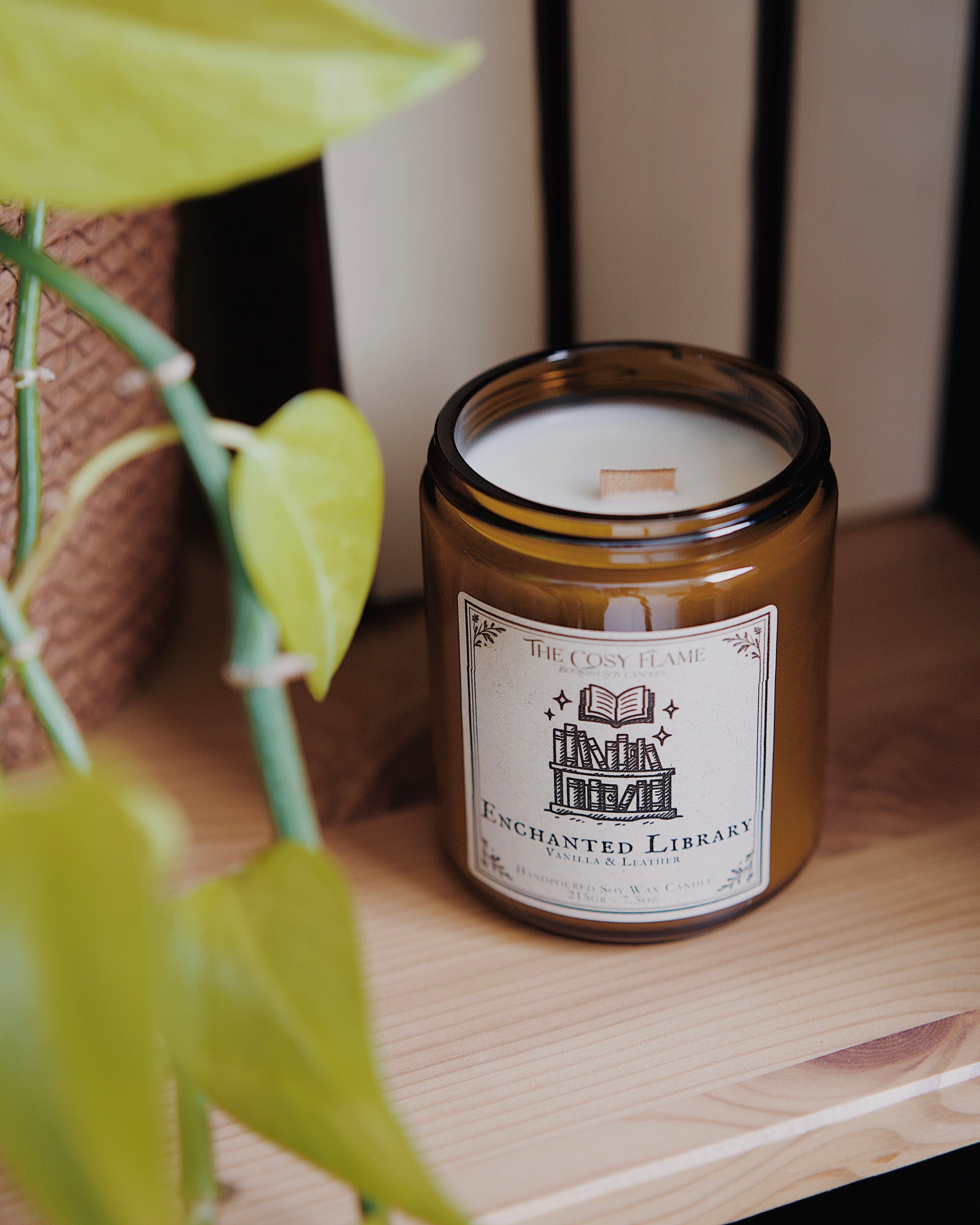 Enchanted Library soy candle by The Cosy Flame in an amber glass jar with a wooden wick, placed on a wooden shelf. Label features a library illustration and featuring the scents vanilla and leather.