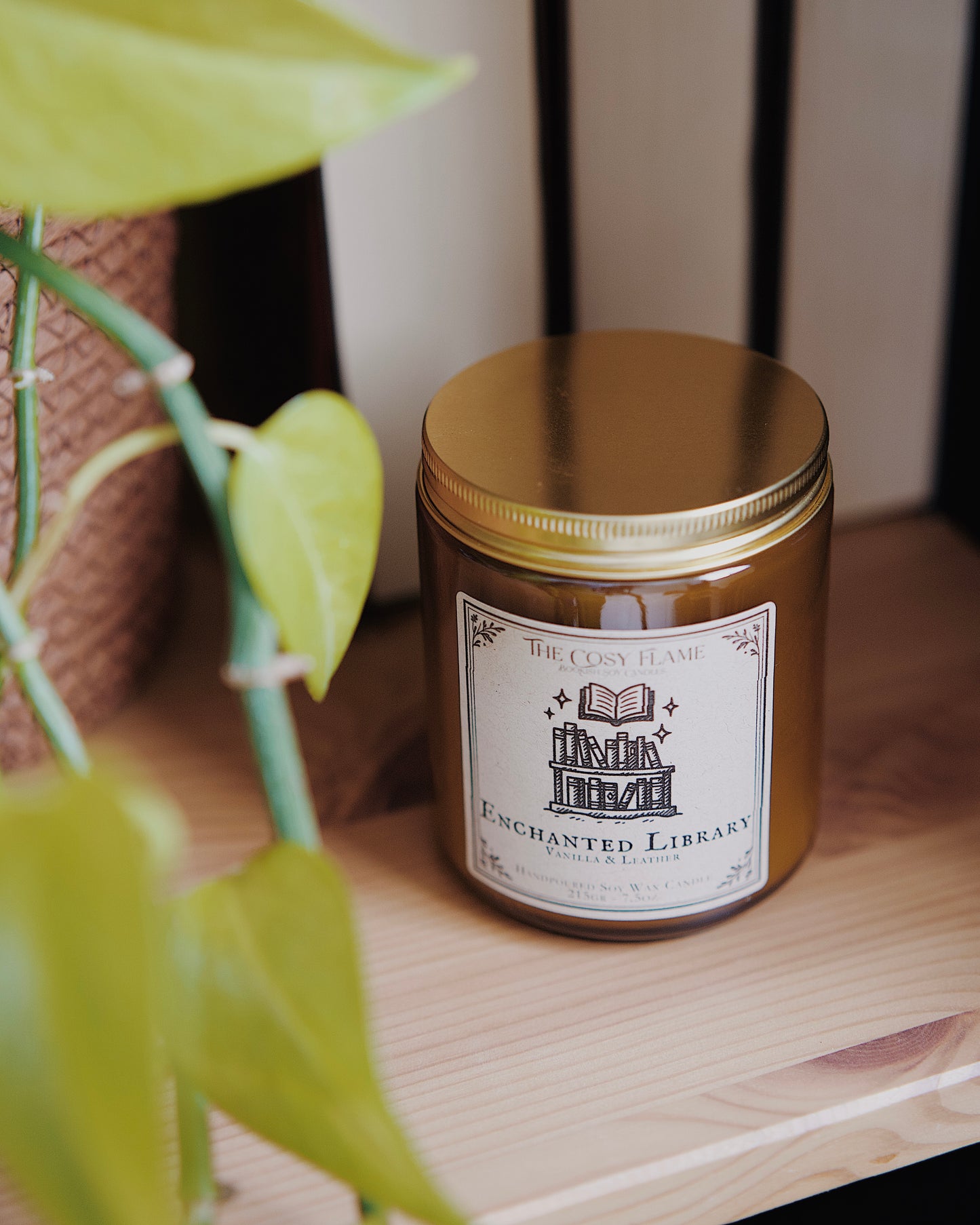 Enchanted Library soy candle by The Cosy Flame in an amber glass jar with a golden lid, placed on a wooden shelf. Label features a library illustration and featuring the scents vanilla and leather.