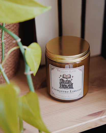 Enchanted Library soy candle by The Cosy Flame in an amber glass jar with a golden lid, placed on a wooden shelf. Label features a library illustration and featuring the scents vanilla and leather.