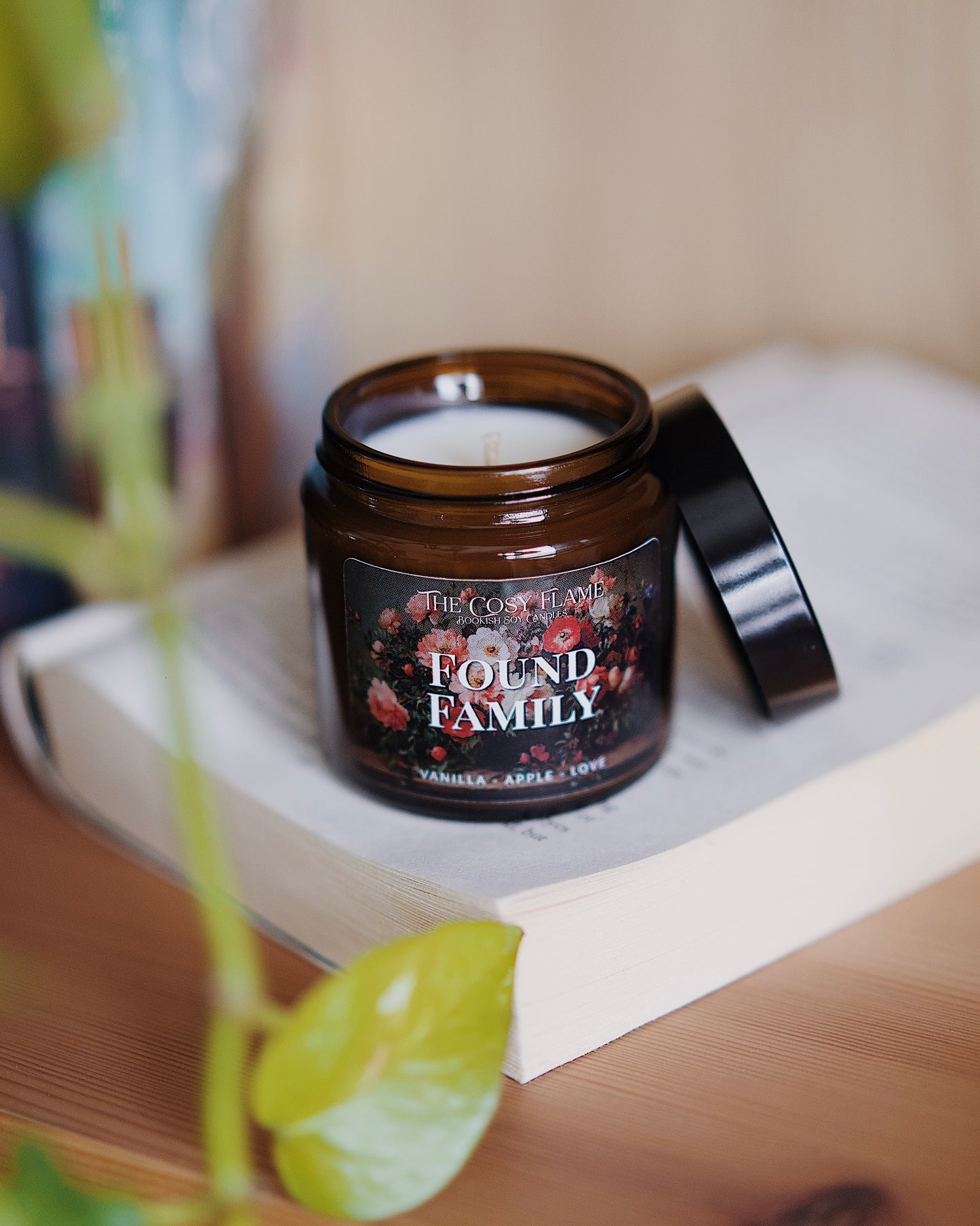 Amber glass candle jar labelled ‘Found Family’ from The Cosy Flame, scented with vanilla and apple, and books in the background.