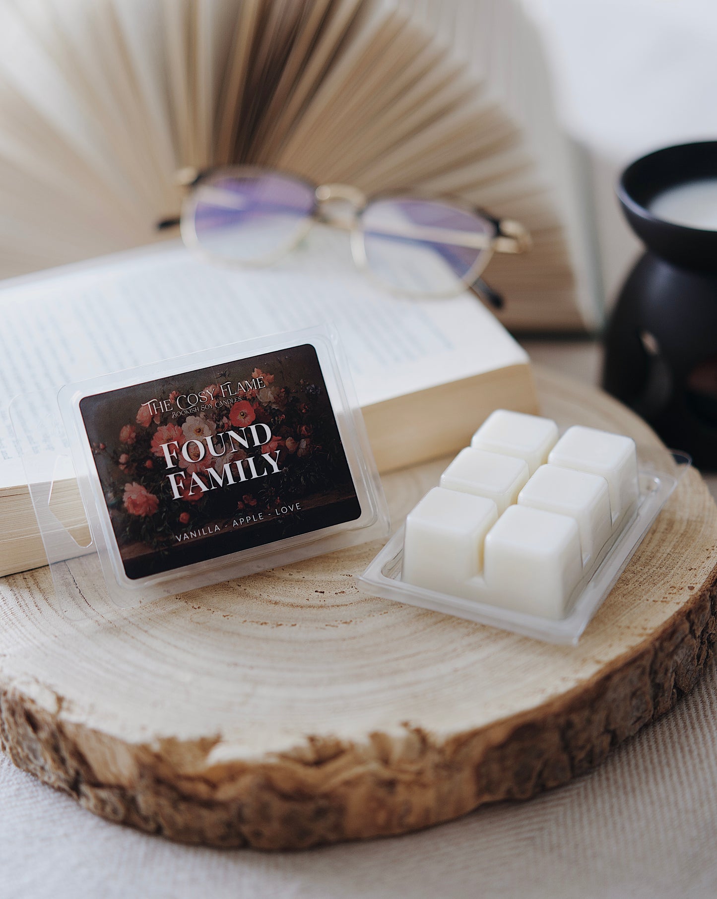 Found Family soy wax melts by The Cosy Flame, featuring vanilla and apple scent notes, displayed on a wooden slice beside an open book and glasses.