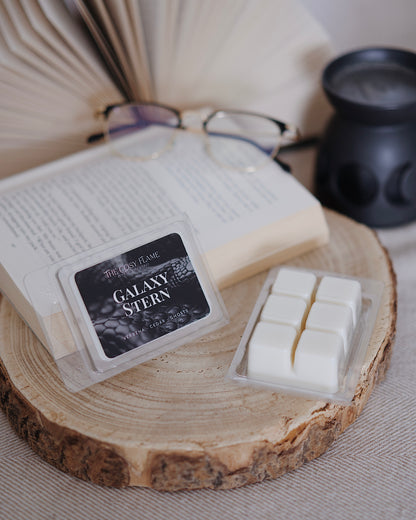 Galaxy Stern soy wax melts by The Cosy Flame, featuring verbena and cedar scent notes, displayed on a wooden slice beside an open book and glasses.