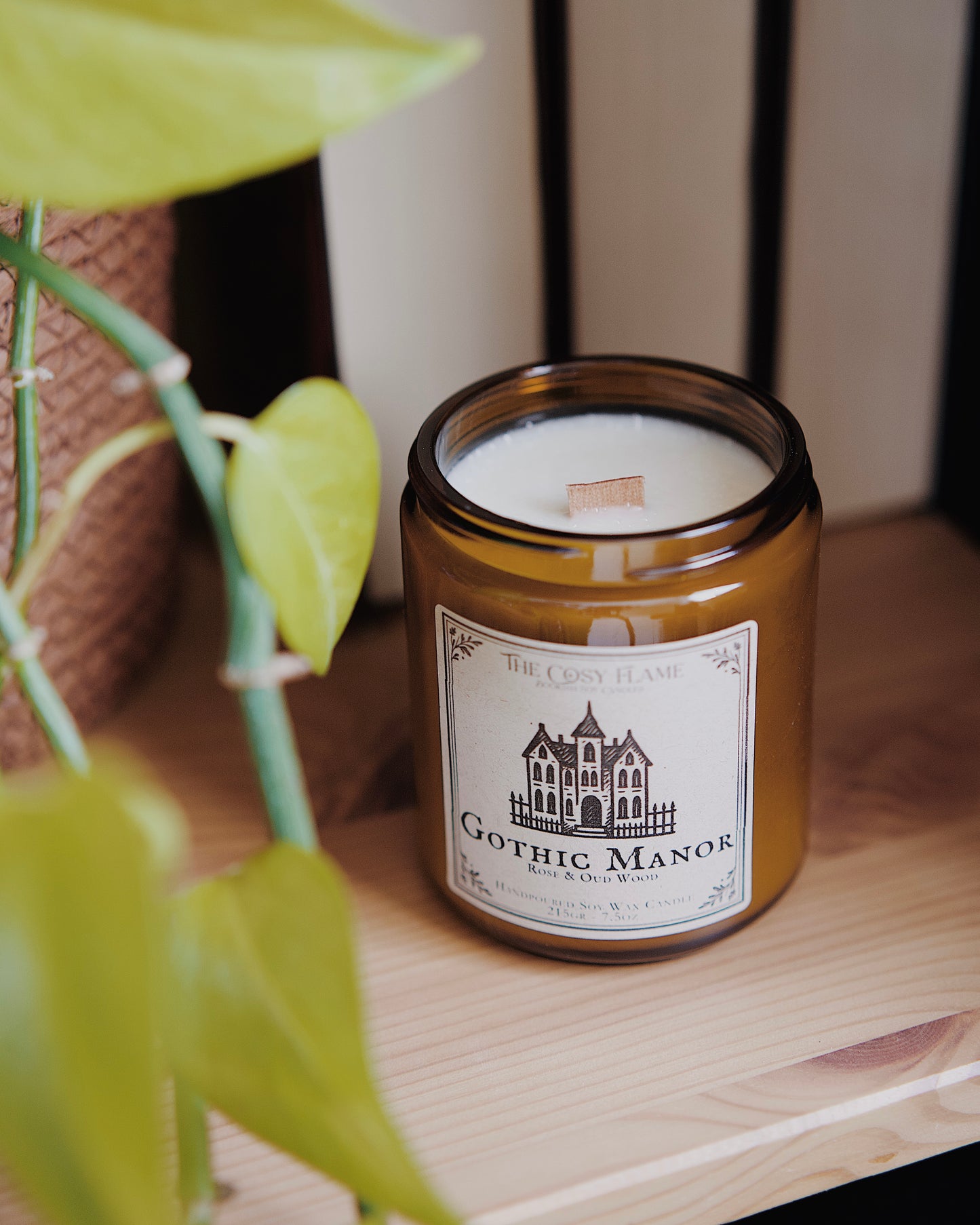 Gothic Manor soy candle by The Cosy Flame in an amber glass jar with a wooden wick, placed on a wooden shelf. Label features a gothic manor illustration and featuring the scents rose and oud wood.