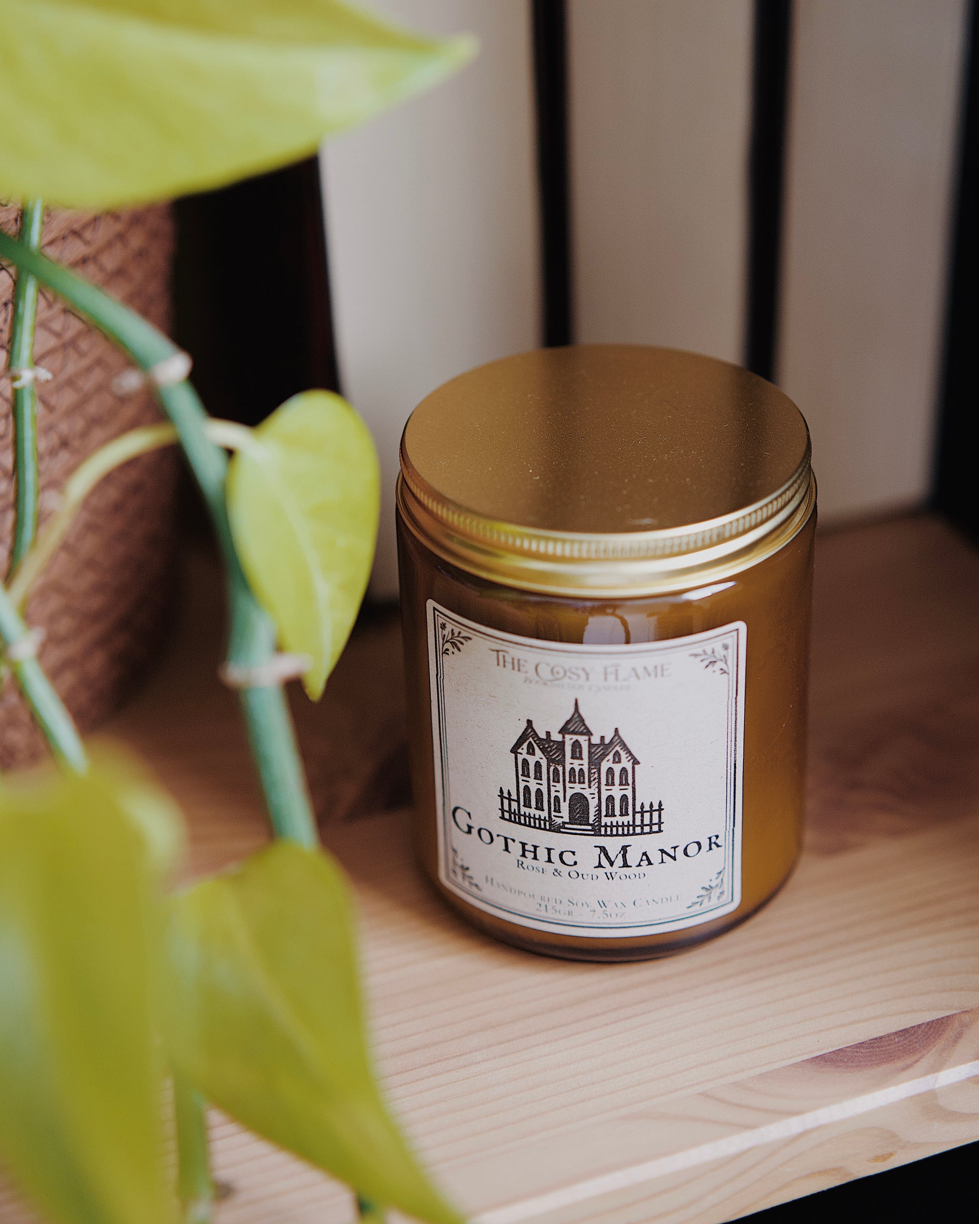 Gothic Manor soy candle by The Cosy Flame in an amber glass jar with a golden lid, placed on a wooden shelf. Label features a gothic manor illustration and featuring the scents rose and oud wood.