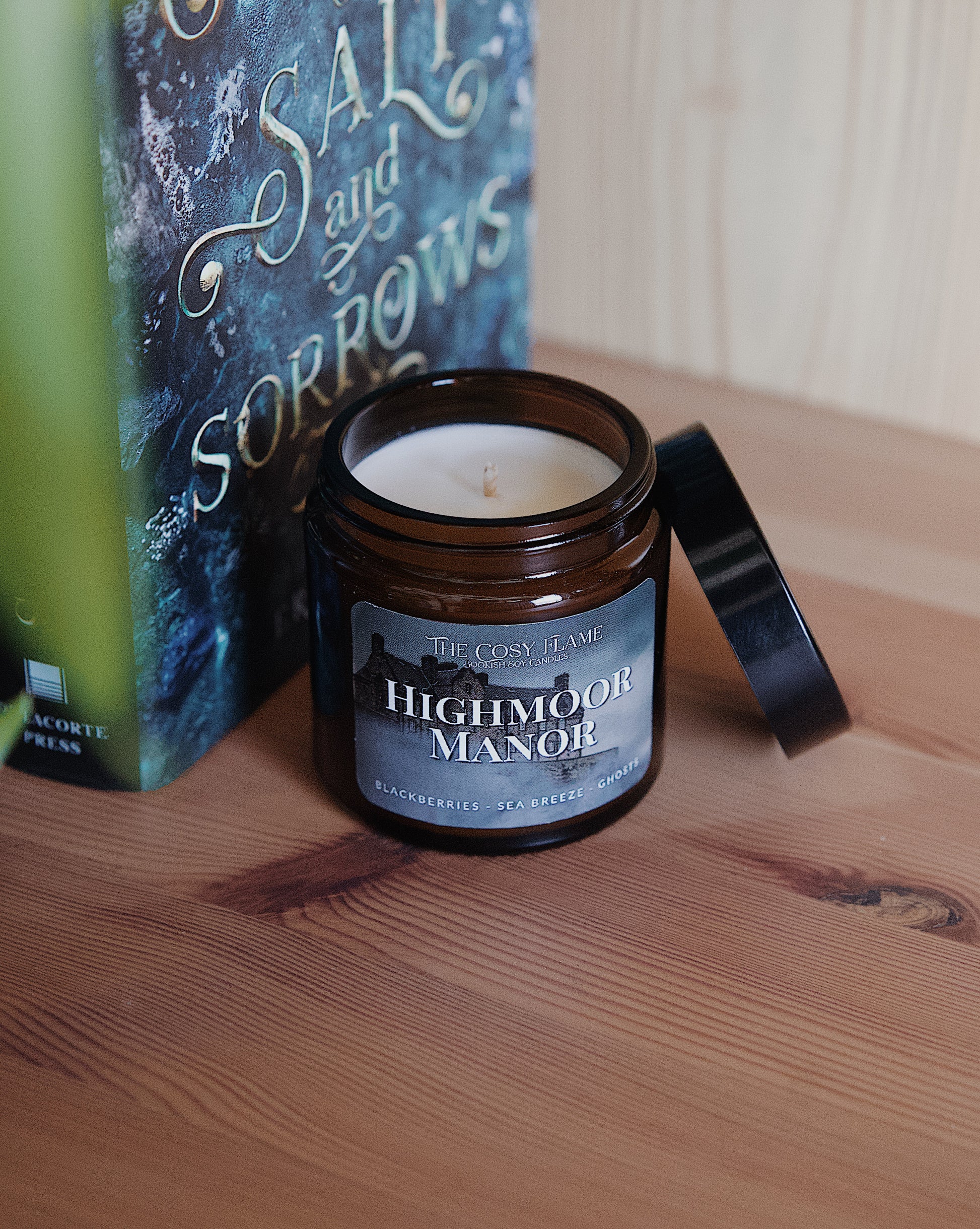 Amber glass candle jar labelled ‘Highmoor Manor’ from The Cosy Flame, scented with blackberries and sea breeze, and House of Salt and Sorrows book in the background.