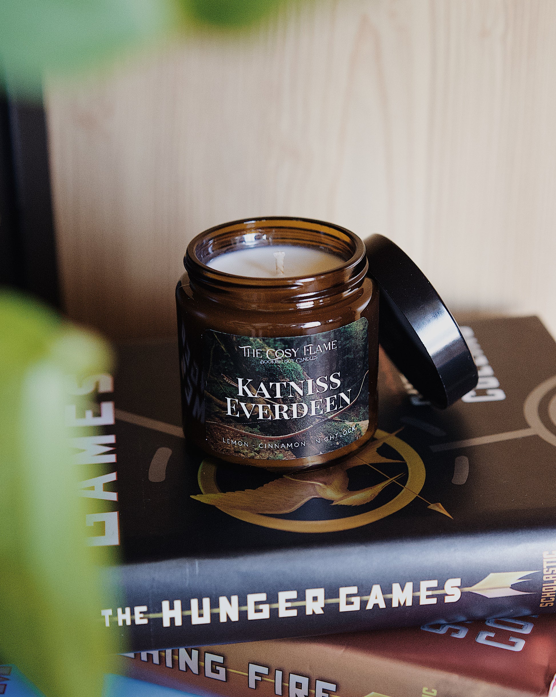 Amber glass candle jar labelled ‘Katniss Everdeen’ from The Cosy Flame, scented with lemon and cinnamon, and The Hunger Games books in the background.