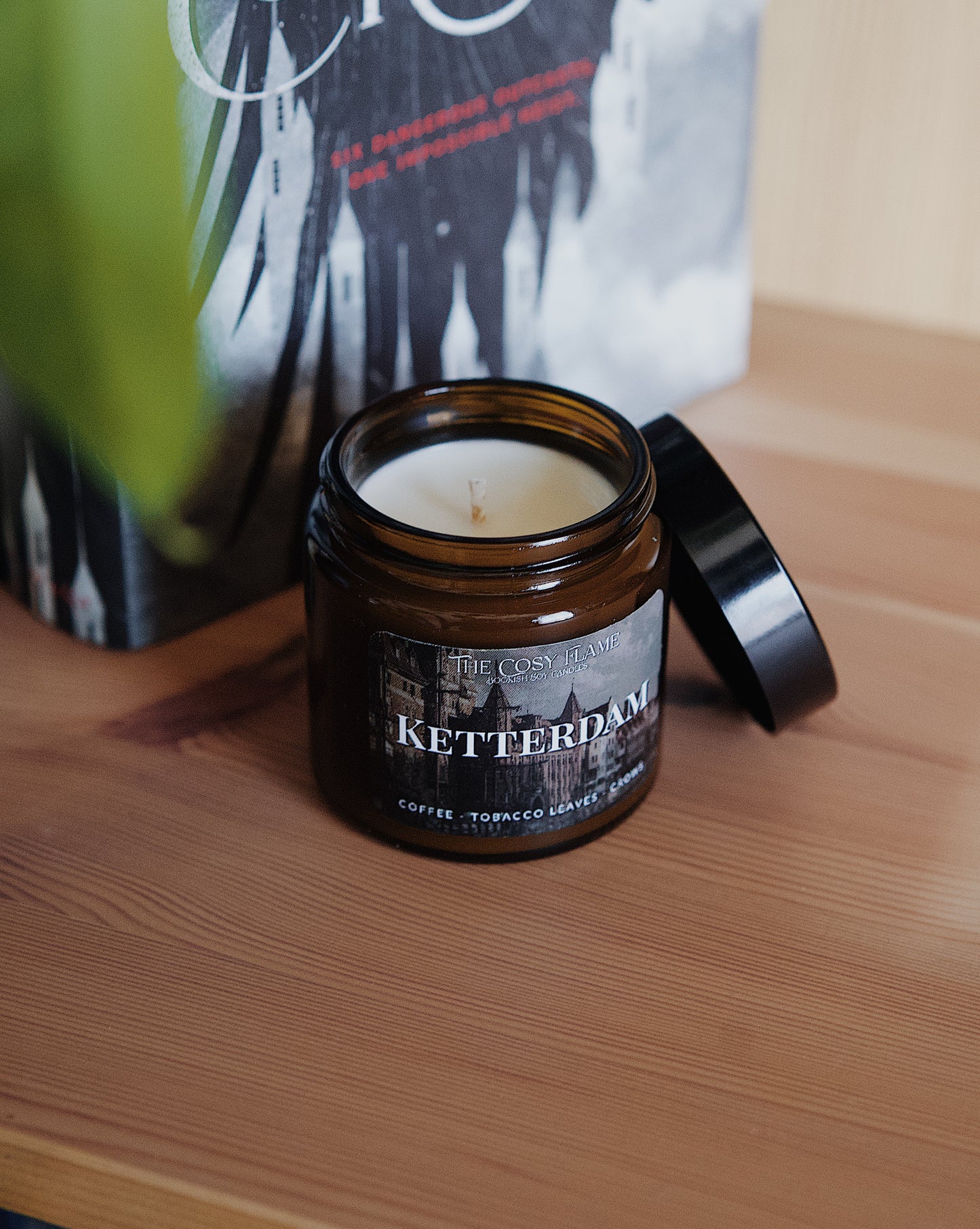 Amber glass candle jar labelled ‘Ketterdam’ from The Cosy Flame, scented with coffee and tobacco leaves, and Six of Crows book in the background.