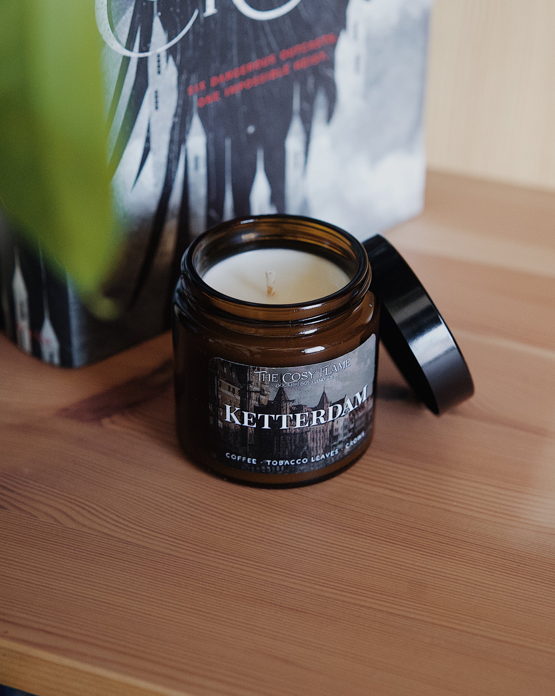 Amber glass candle jar labelled ‘Ketterdam’ from The Cosy Flame, scented with coffee and tobacco leaves, and Six of Crows book in the background.