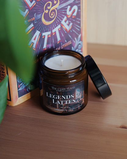 Amber glass candle jar labelled ‘Legends & Lattes’ from The Cosy Flame, scented with coffee and cardamom, and Legends & Lattes book in the background.
