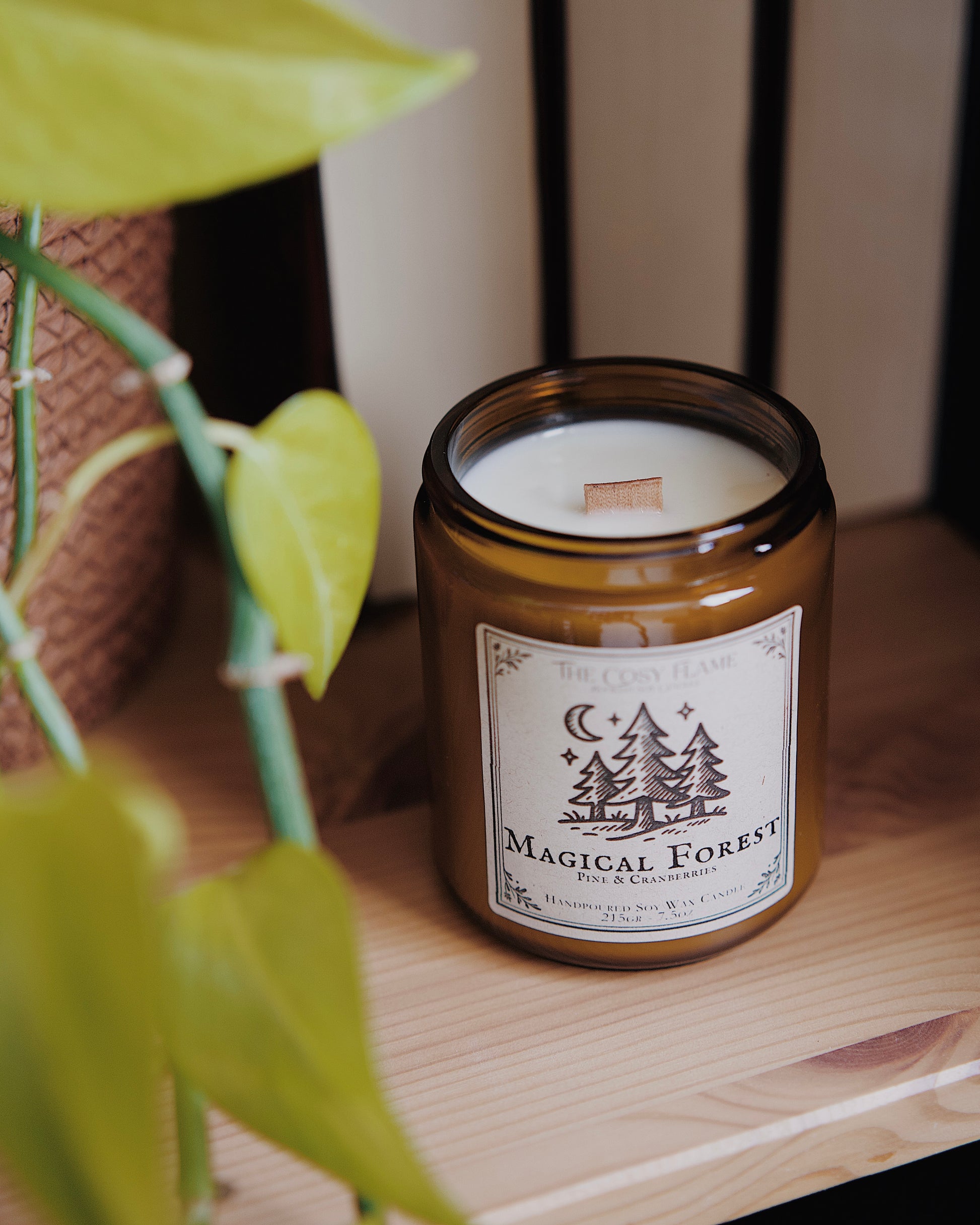 Magical Forest soy candle by The Cosy Flame in an amber glass jar with a wooden wick, placed on a wooden shelf. Label features a forest in the moonlight illustration and featuring the scents pine and cranberries.