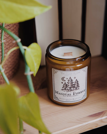 Magical Forest soy candle by The Cosy Flame in an amber glass jar with a wooden wick, placed on a wooden shelf. Label features a forest in the moonlight illustration and featuring the scents pine and cranberries.