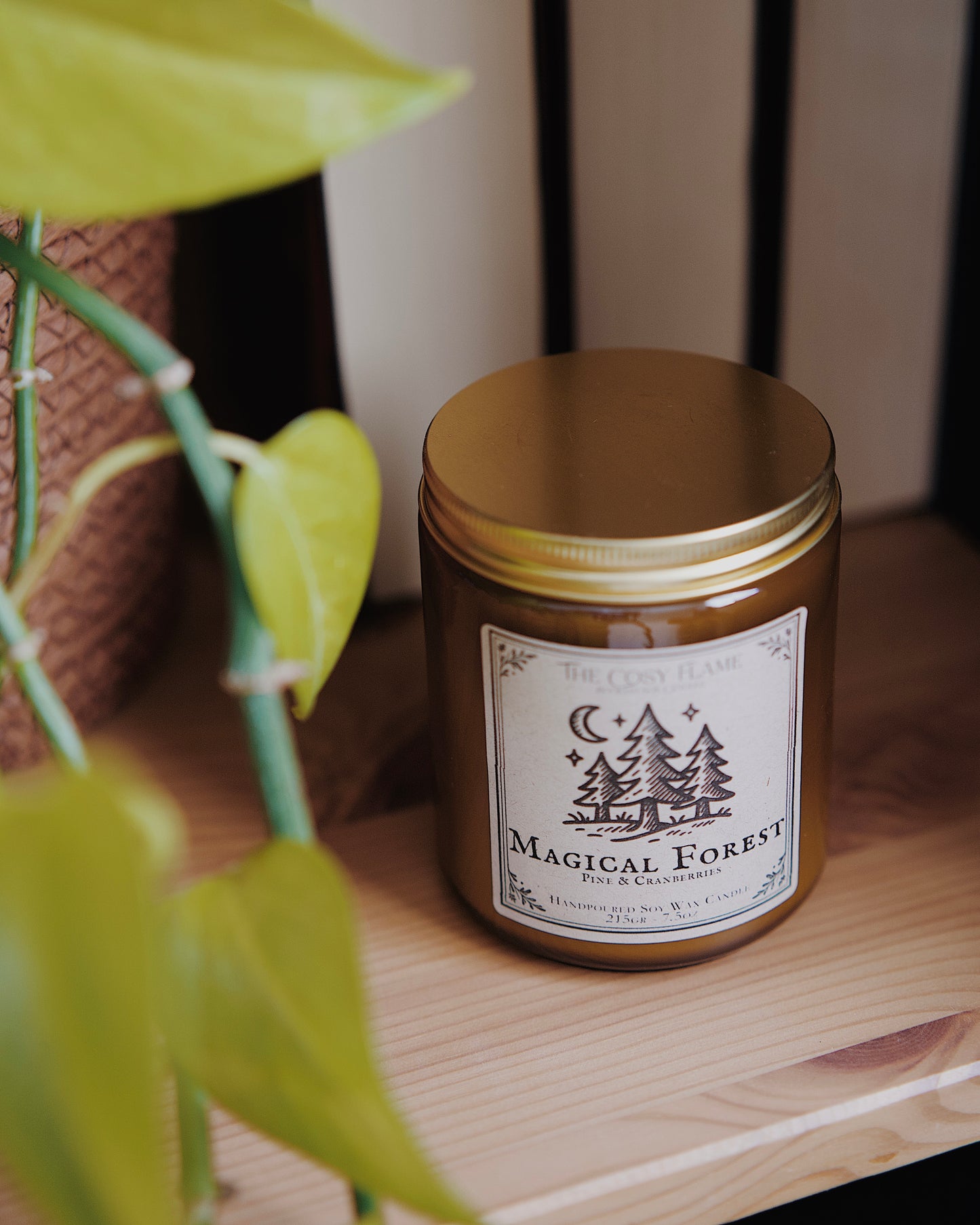Magical Forest soy candle by The Cosy Flame in an amber glass jar with a golden lid, placed on a wooden shelf. Label features a forest in the moonlight illustration and featuring the scents pine and cranberries.