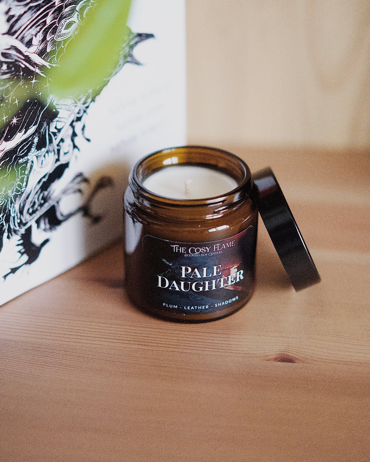 Amber glass candle jar labelled ‘Pale Daughter’ from The Cosy Flame, scented with plum and leather, and Nevernight book in the background.