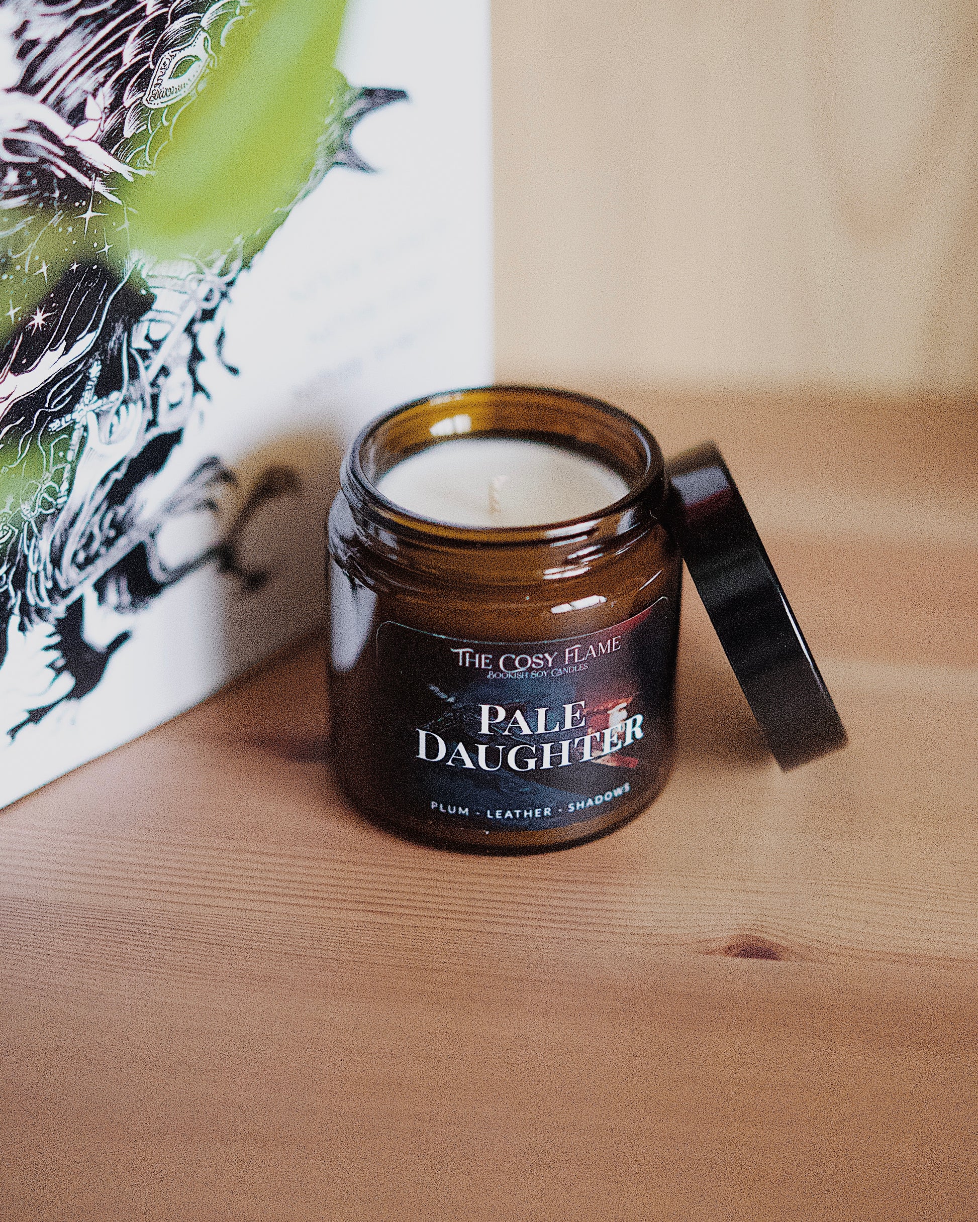 Amber glass candle jar labelled ‘Pale Daughter’ from The Cosy Flame, scented with plum and leather, and Nevernight book in the background.