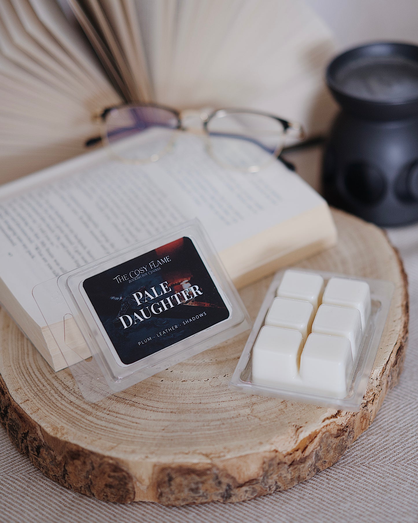 Pale Daughter soy wax melts by The Cosy Flame, featuring plum and leather scent notes, displayed on a wooden slice beside an open book and glasses.