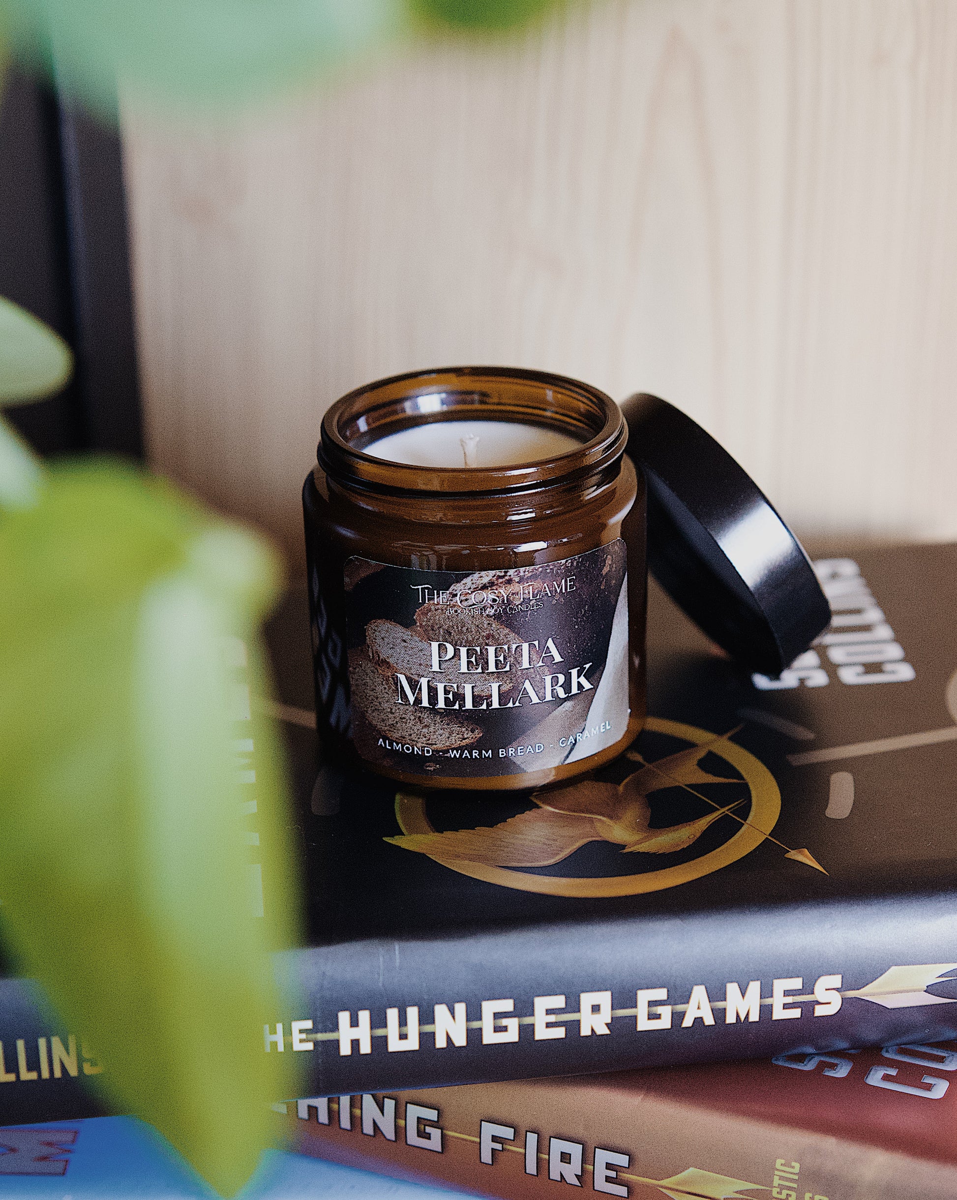 Amber glass candle jar labelled ‘Peeta Mellark’ from The Cosy Flame, scented with almond, warm bread and caramel, and The Hunger Games books in the background.
