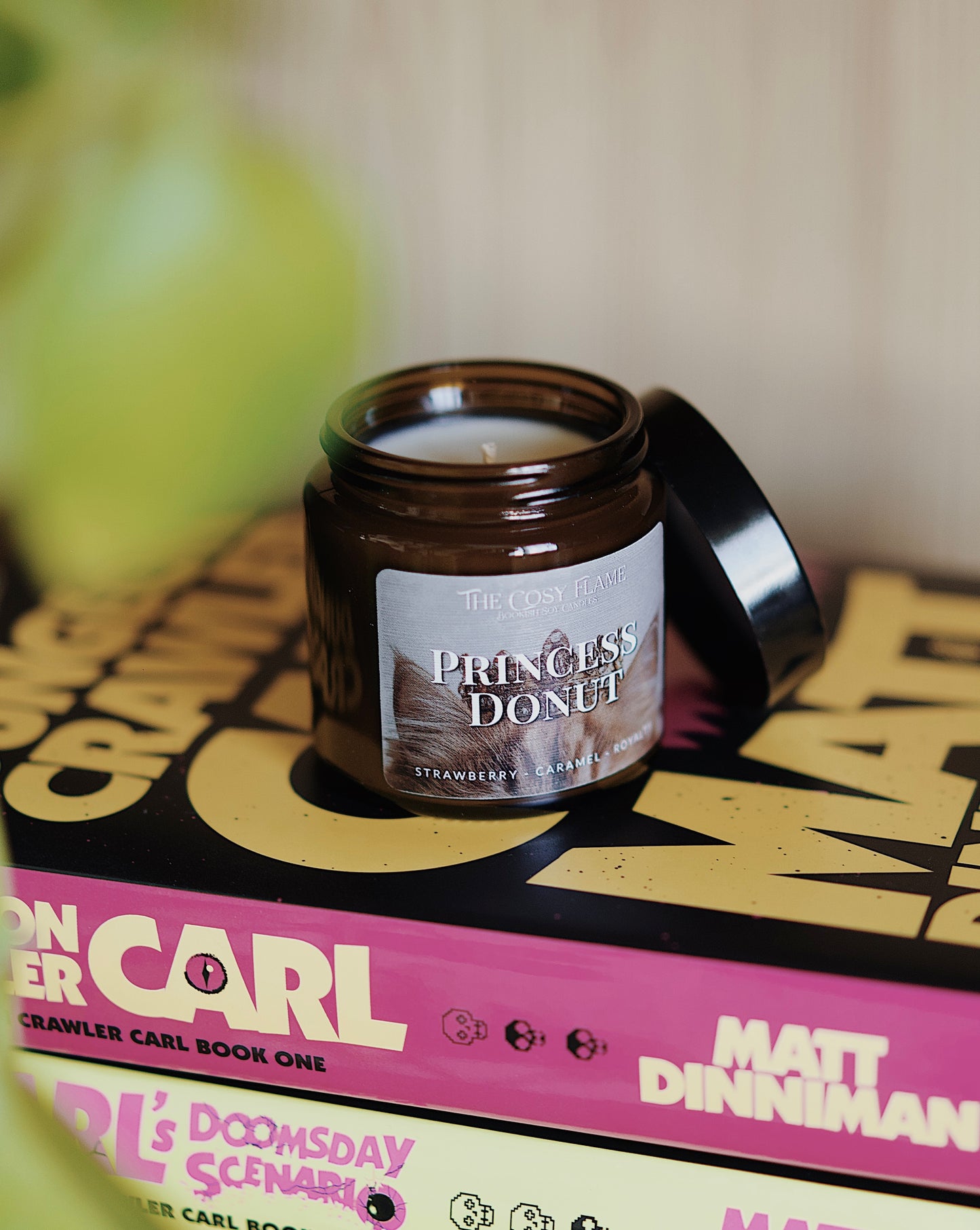 Amber glass candle jar labelled ‘Princess Donut’ from The Cosy Flame, scented with strawberry and caramel, and Dungeon Crawler Carl books in the background.