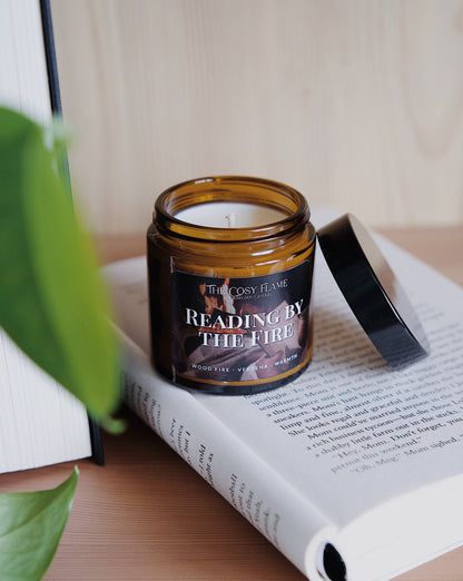 Amber glass candle jar labelled ‘Reading by the Fire’ from The Cosy Flame, scented with wood fire and verbena, and book in the background.