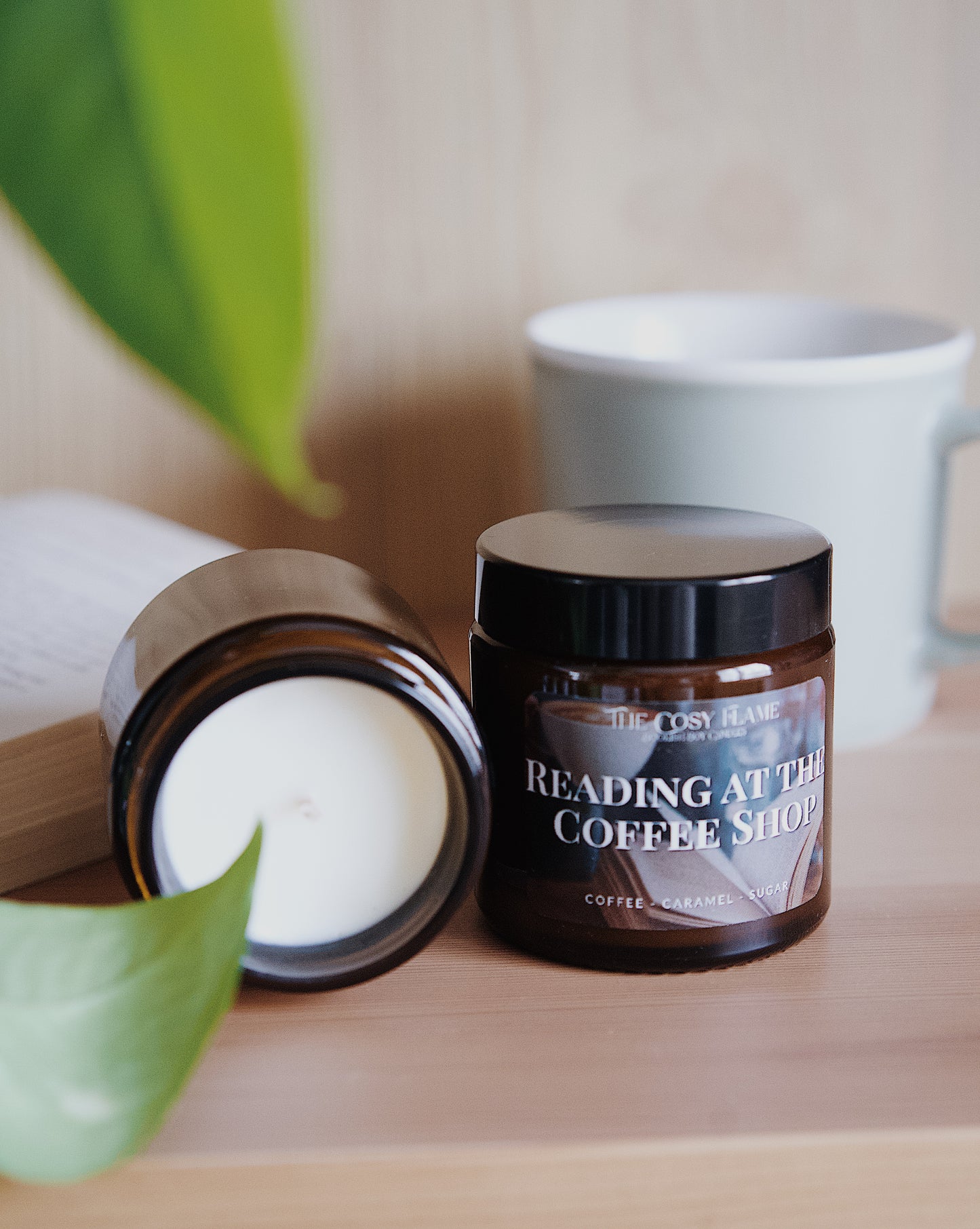 Amber glass candle jar labelled ‘Reading at the Coffee Shop’ from The Cosy Flame, scented with coffee and caramel, and book and coffee mug in the background.