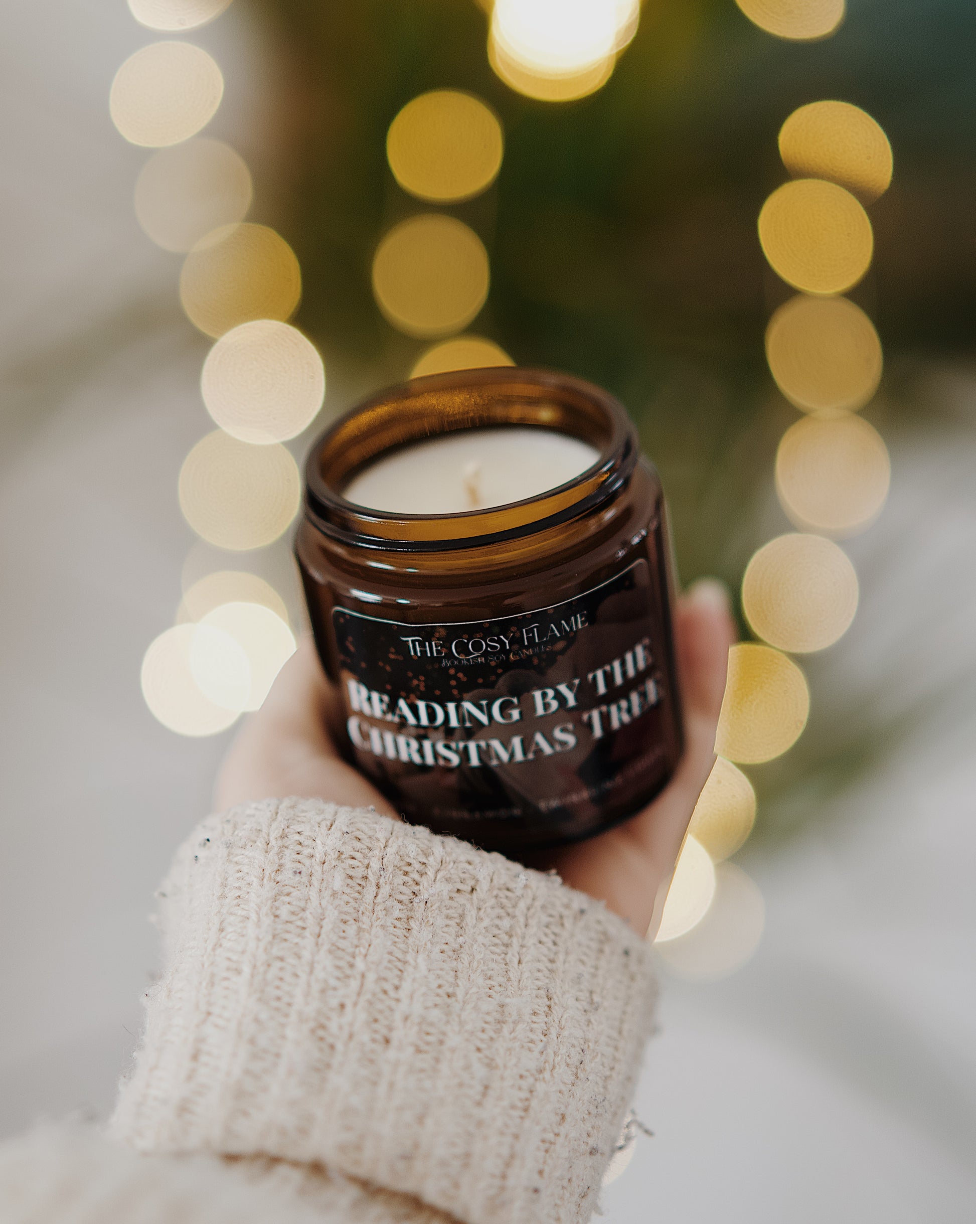 Amber glass candle jar labelled ‘Reading by the Christmas Tree’ from The Cosy Flame, scented with orange and cinnamon, and Christmas tree in the background.