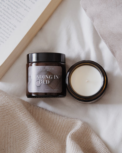 Amber glass candle jar labelled ‘Reading in Bed’ from The Cosy Flame, scented with cotton, peach and fresh laundry, and book on bed in the background.