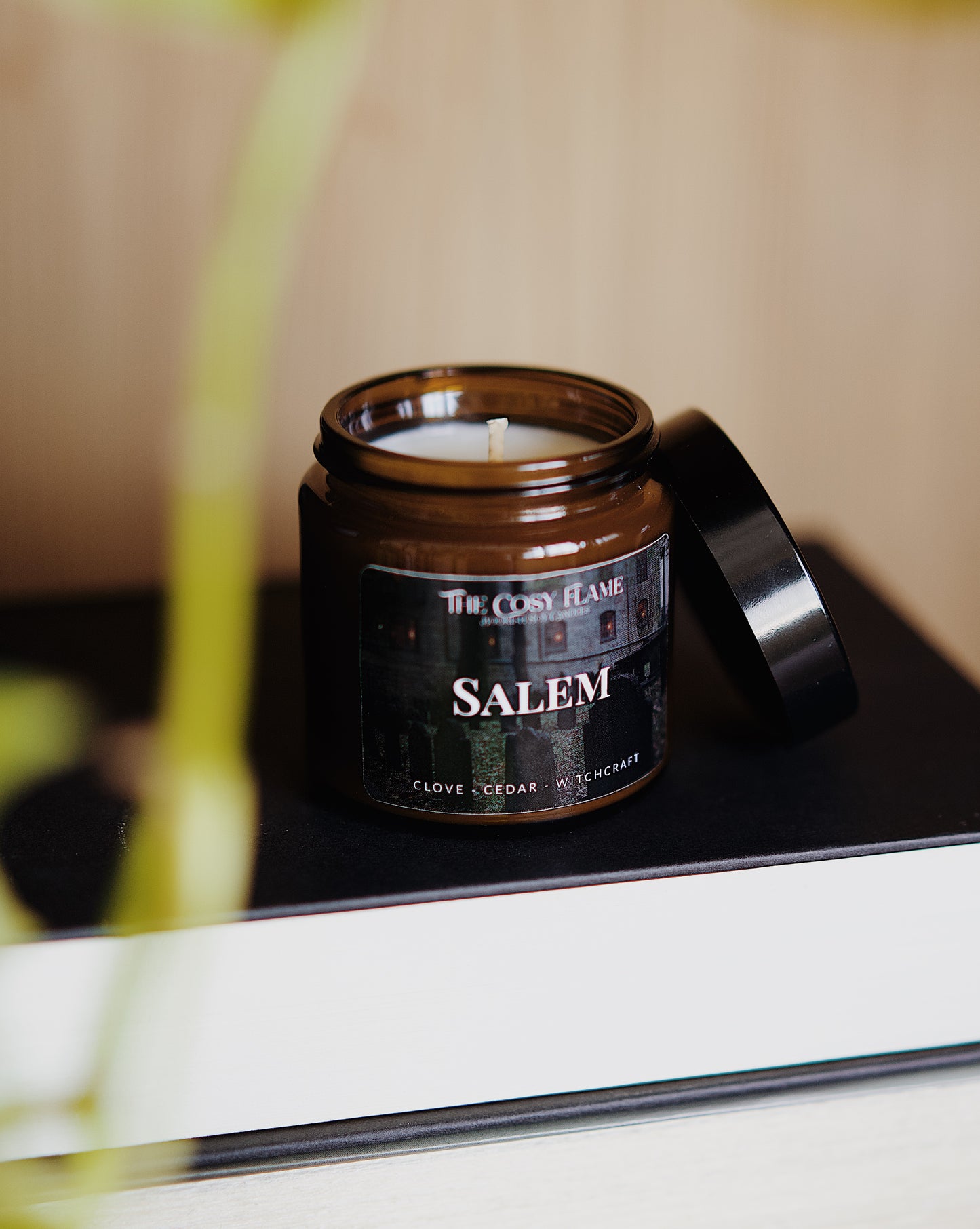 Amber glass candle jar labelled ‘Salem’ from The Cosy Flame, scented with clove and cedar, and books in the background.