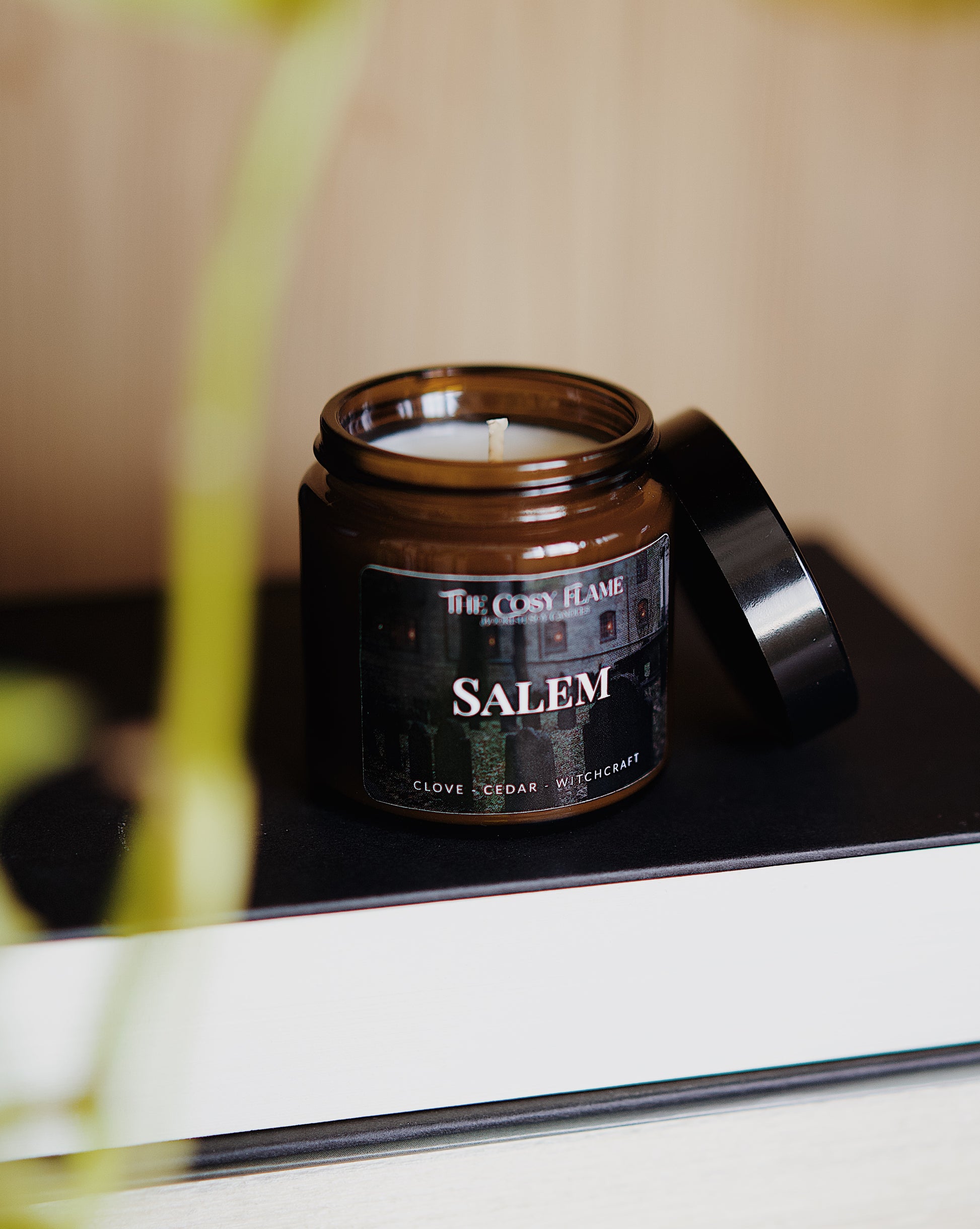 Amber glass candle jar labelled ‘Salem’ from The Cosy Flame, scented with clove and cedar, and books in the background.