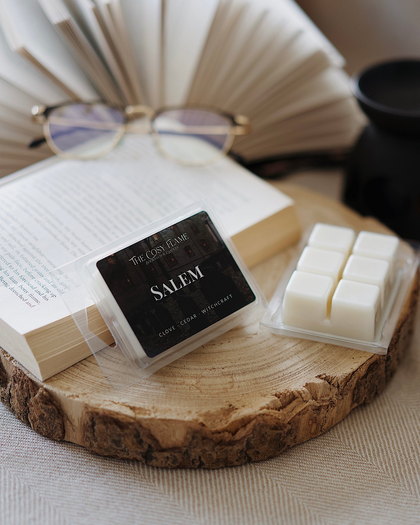 Salem soy wax melts by The Cosy Flame, featuring clove and cedar scent notes, displayed on a wooden slice beside an open book and glasses.