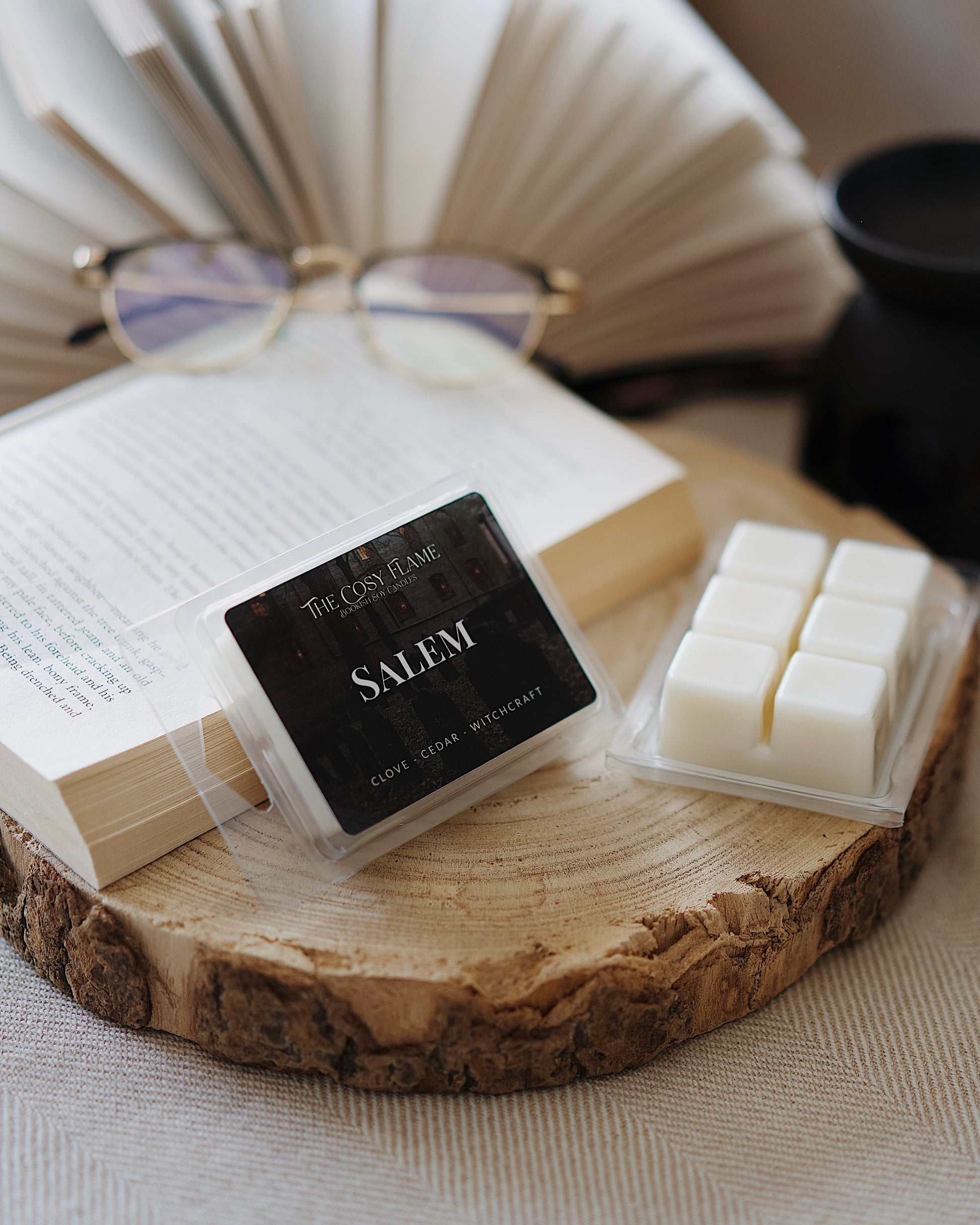 Salem soy wax melts by The Cosy Flame, featuring clove and cedar scent notes, displayed on a wooden slice beside an open book and glasses.