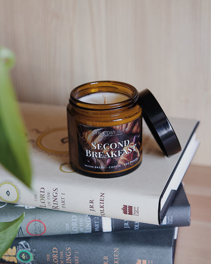 Amber glass candle jar labelled ‘Second Breakfast’ from The Cosy Flame, scented with warm bread, coffee and red fruits, and The Lord of the Rings books in the background.