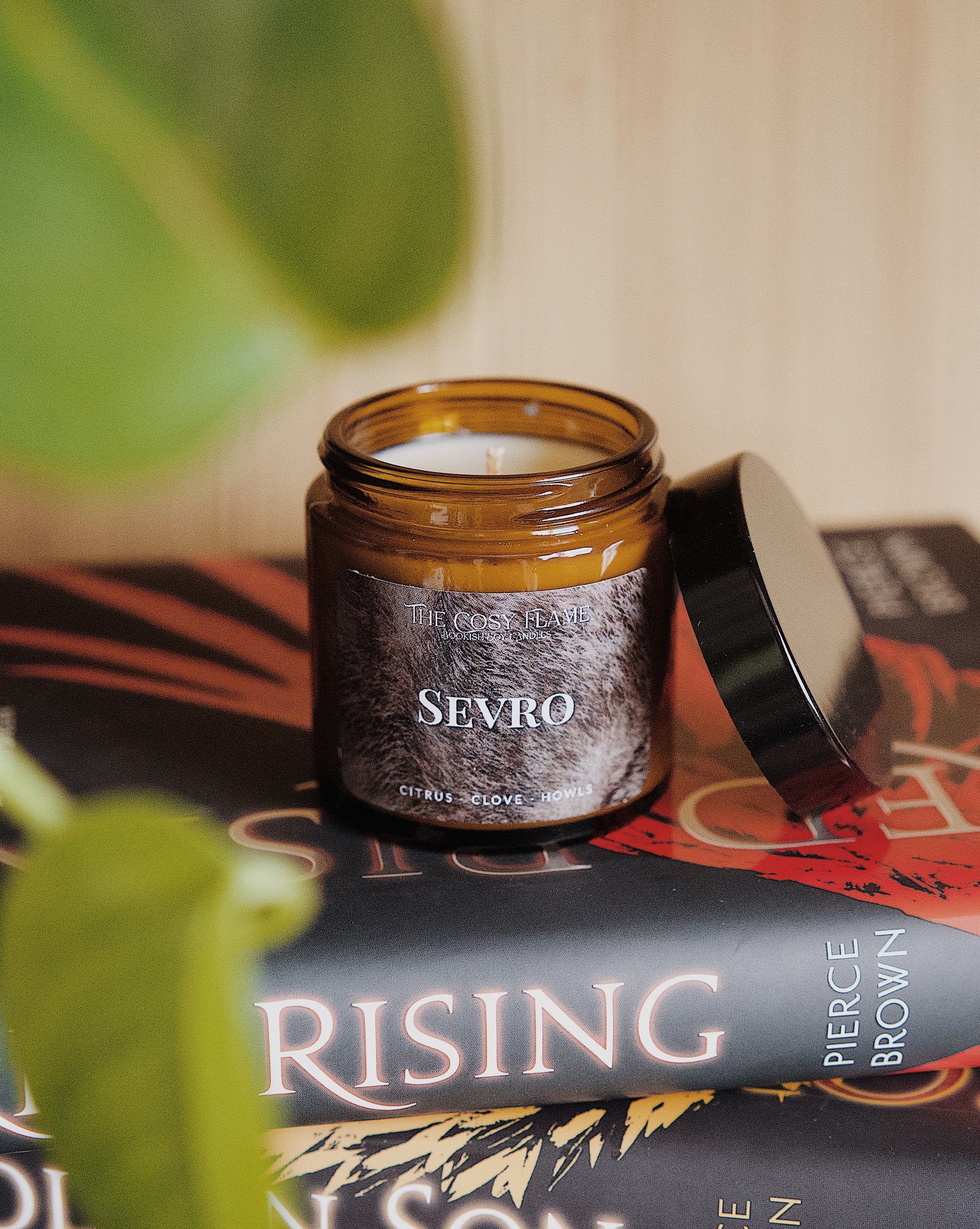 Amber glass candle jar labelled ‘Sevro’ from The Cosy Flame, scented with citrus and clove, and Red Rising books in the background.
