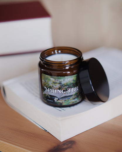 Amber glass candle jar labelled ‘Spring TBR’ from The Cosy Flame, scented with peach, strawberry and flowers, and books in the background.