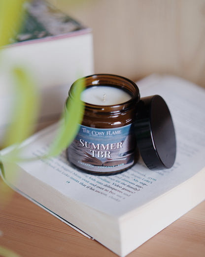 Amber glass candle jar labelled ‘Summer TBR’ from The Cosy Flame, scented with pineapple and coconut, and books in the background.