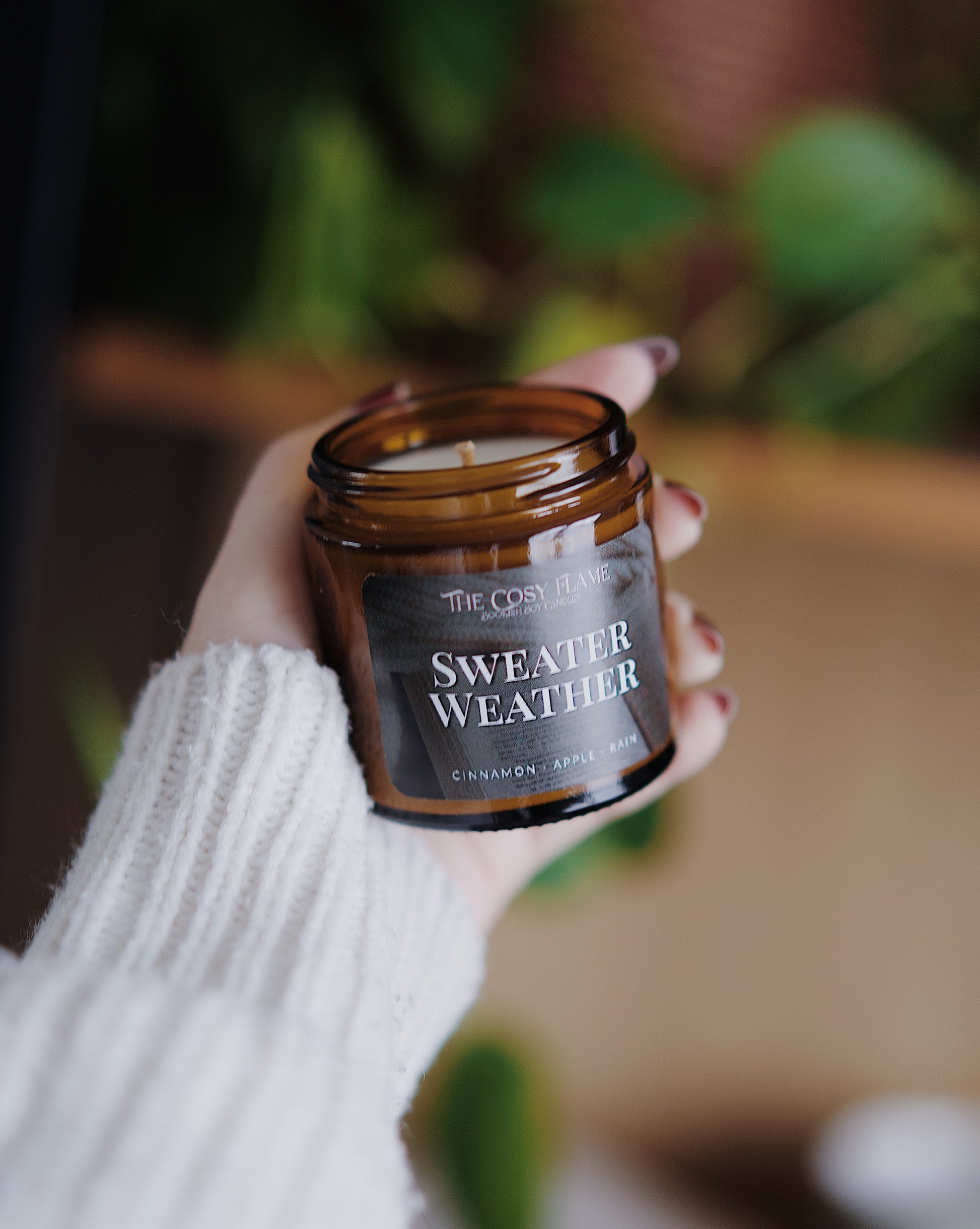 Amber glass candle jar labelled ‘Sweater Weather’ from The Cosy Flame, scented with cinnamon and apple, and book shelves in the background.