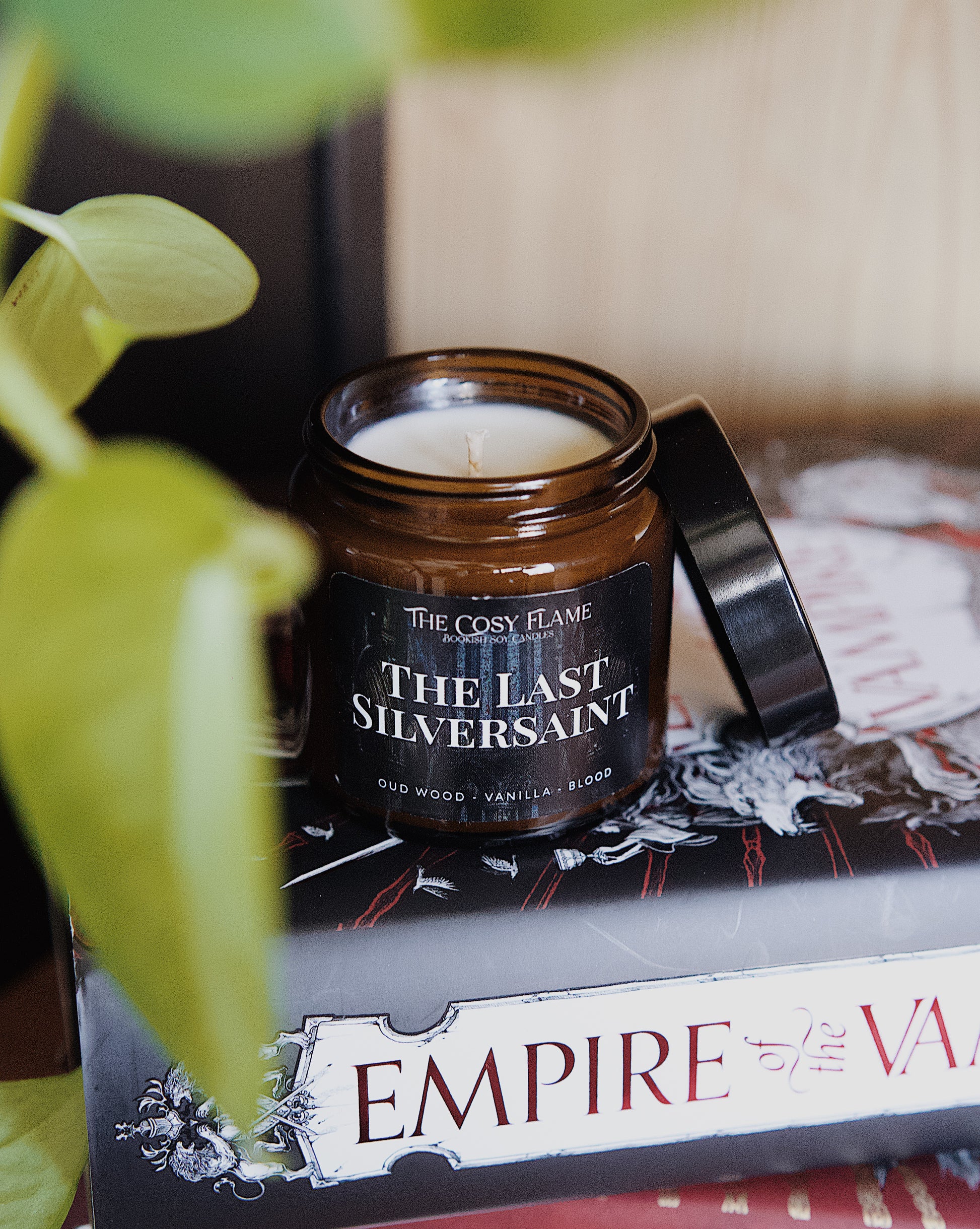Amber glass candle jar labelled ‘The Last Silversaint’ from The Cosy Flame, scented with oud wood and vanilla, and Empire of the Vampire books in the background.